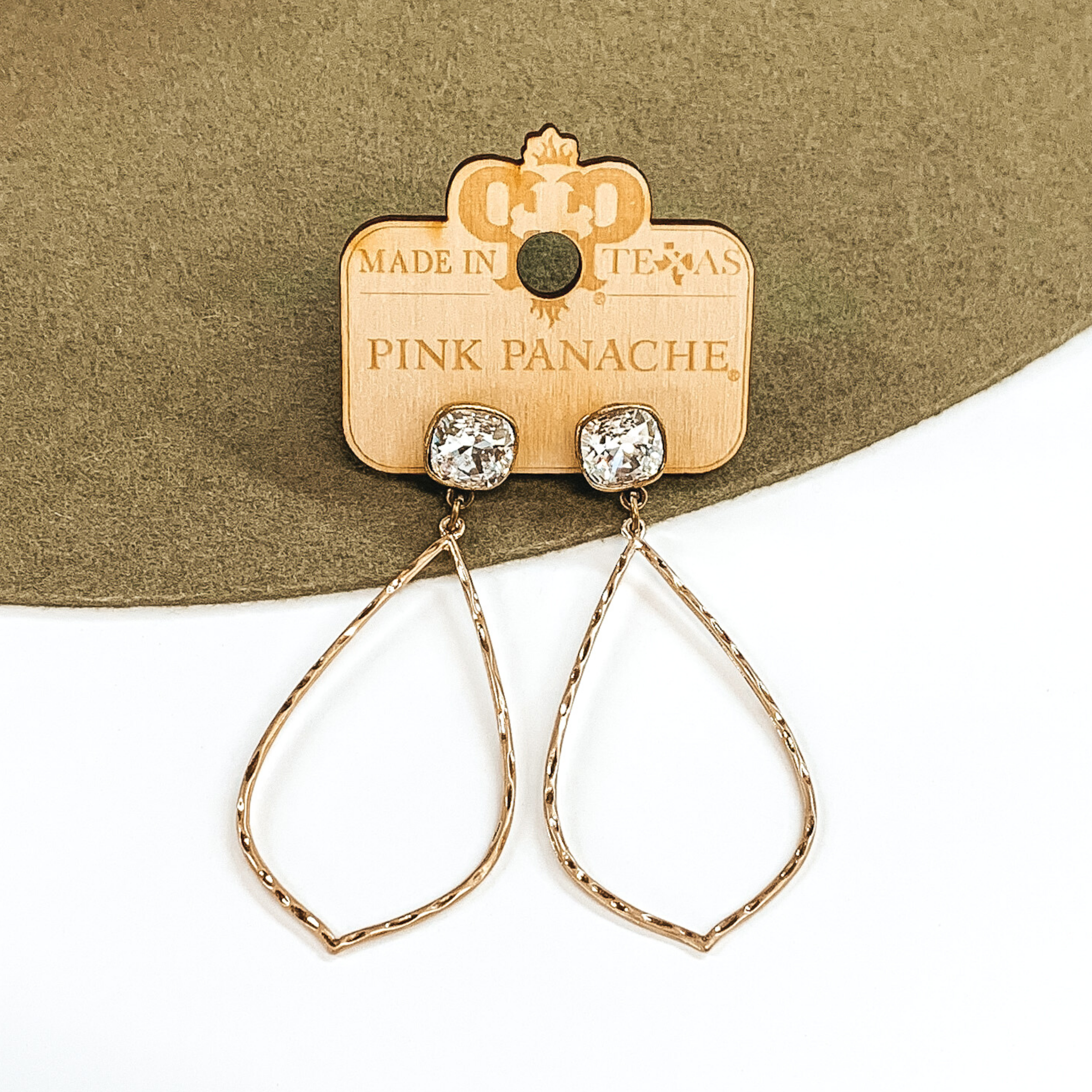 Bronze stud earrings with square clear crystals that has a thin, hammered teardrop outline pendant that hangs from the studs. This pair of earrings are pictured on a white and olive background. 