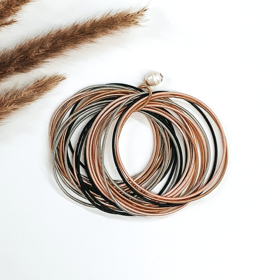 Thing gold, silver, black, and light pink bracelets bunched together with a pearl beaded charm on a white background
