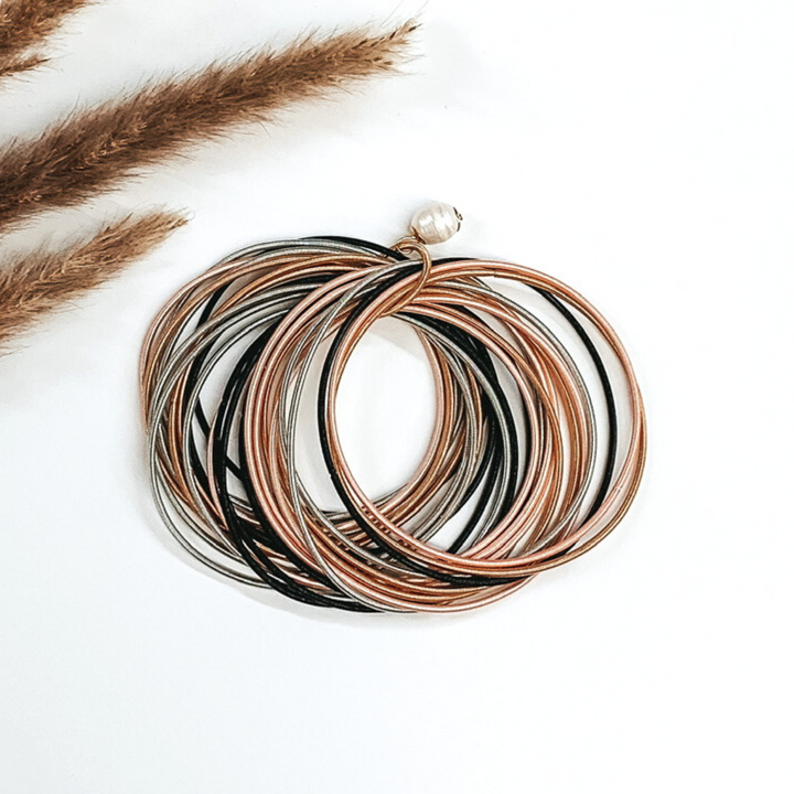 Thing gold, silver, black, and light pink bracelets bunched together with a pearl beaded charm on a white background