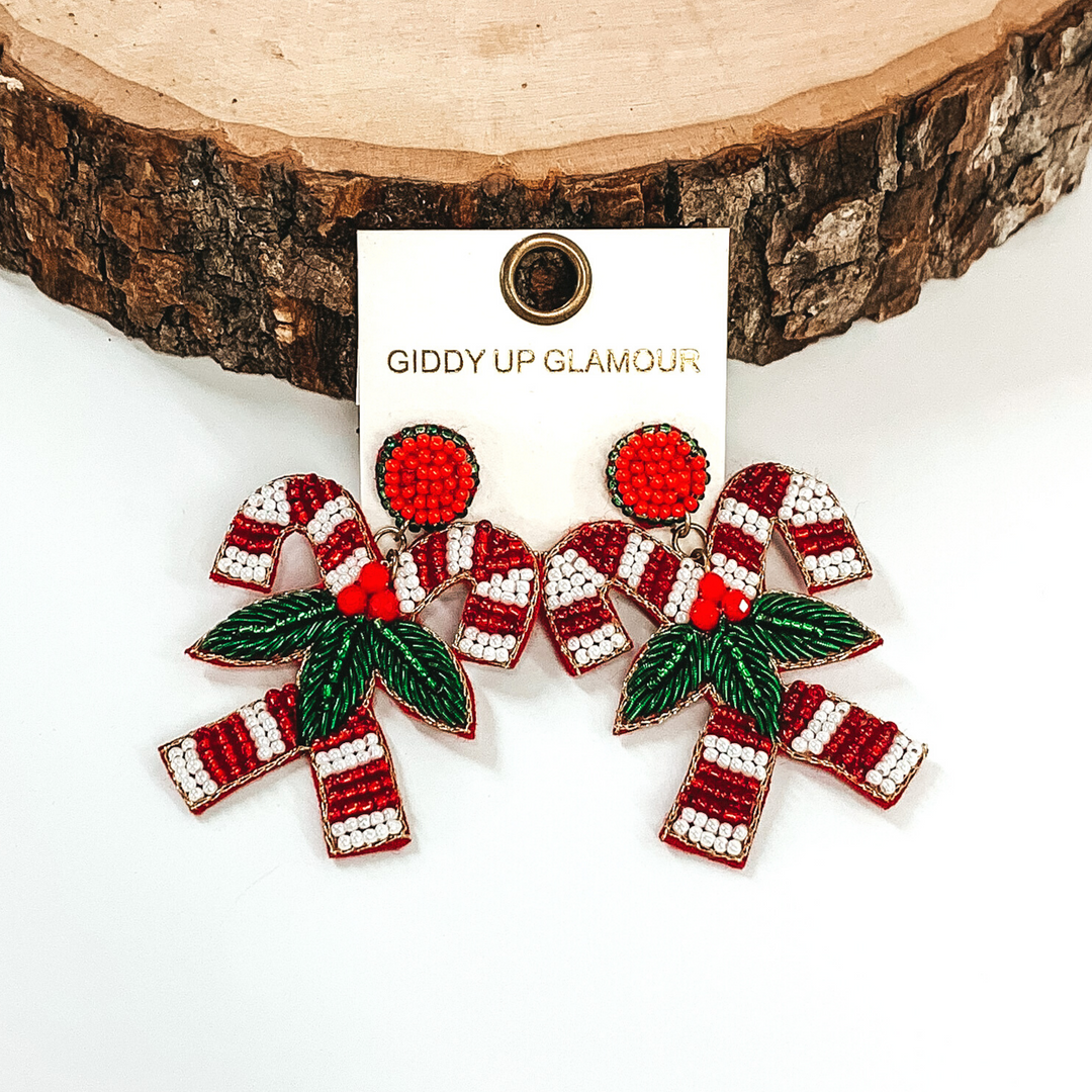 Red and white striped beaded candy canes earrings with them connected together with mistletoe on a white background. 
