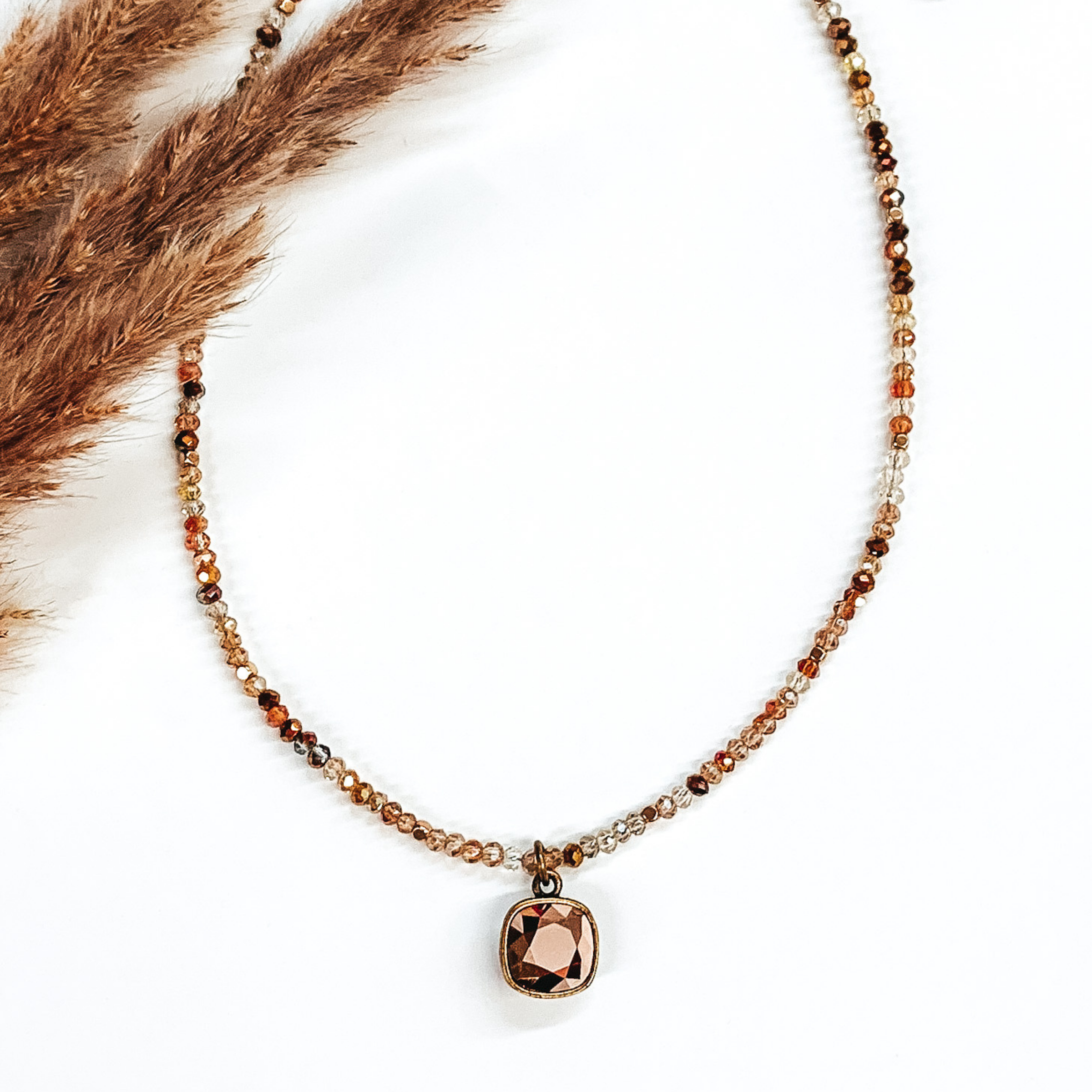 Rose gold and bronze mix beaded adjustable necklace with a single square crystal in a rose gold color. This necklace is pictured of a white background with some brown floral.