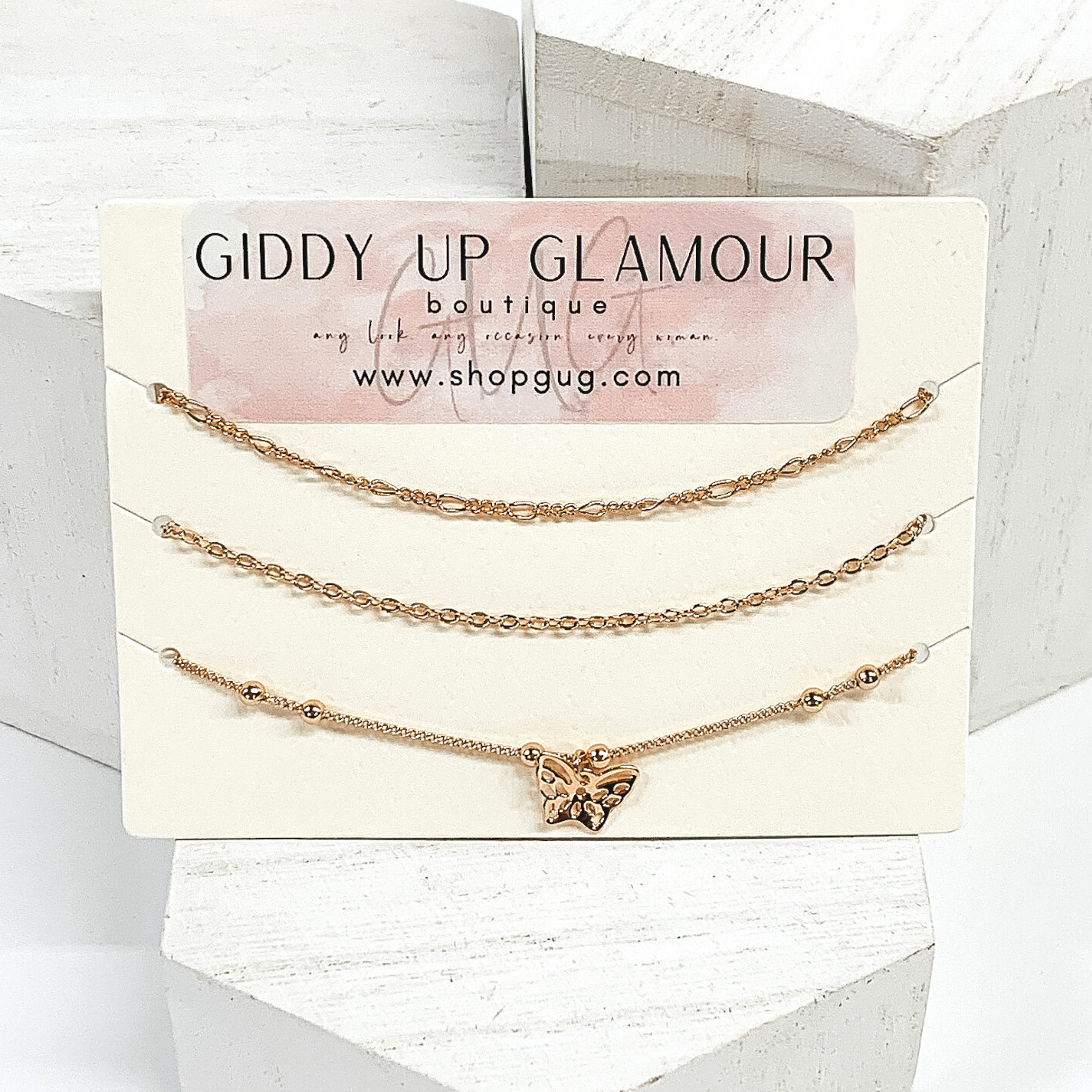 Three gold chained anklets, onw has a single butterfly charm. They are pictured on a white background