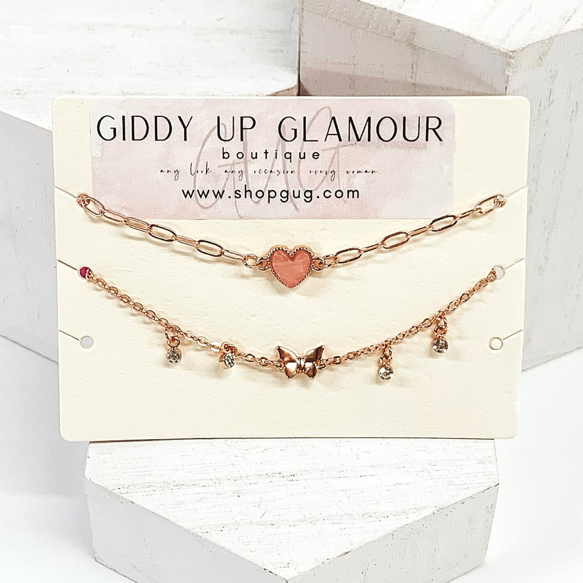 Two gold chained anklets. One anklet has a blush colored heart harm. The second anklet has a small butterfly charm with tiny crystal drop charms surrounding it. These are pictured on a white background. 