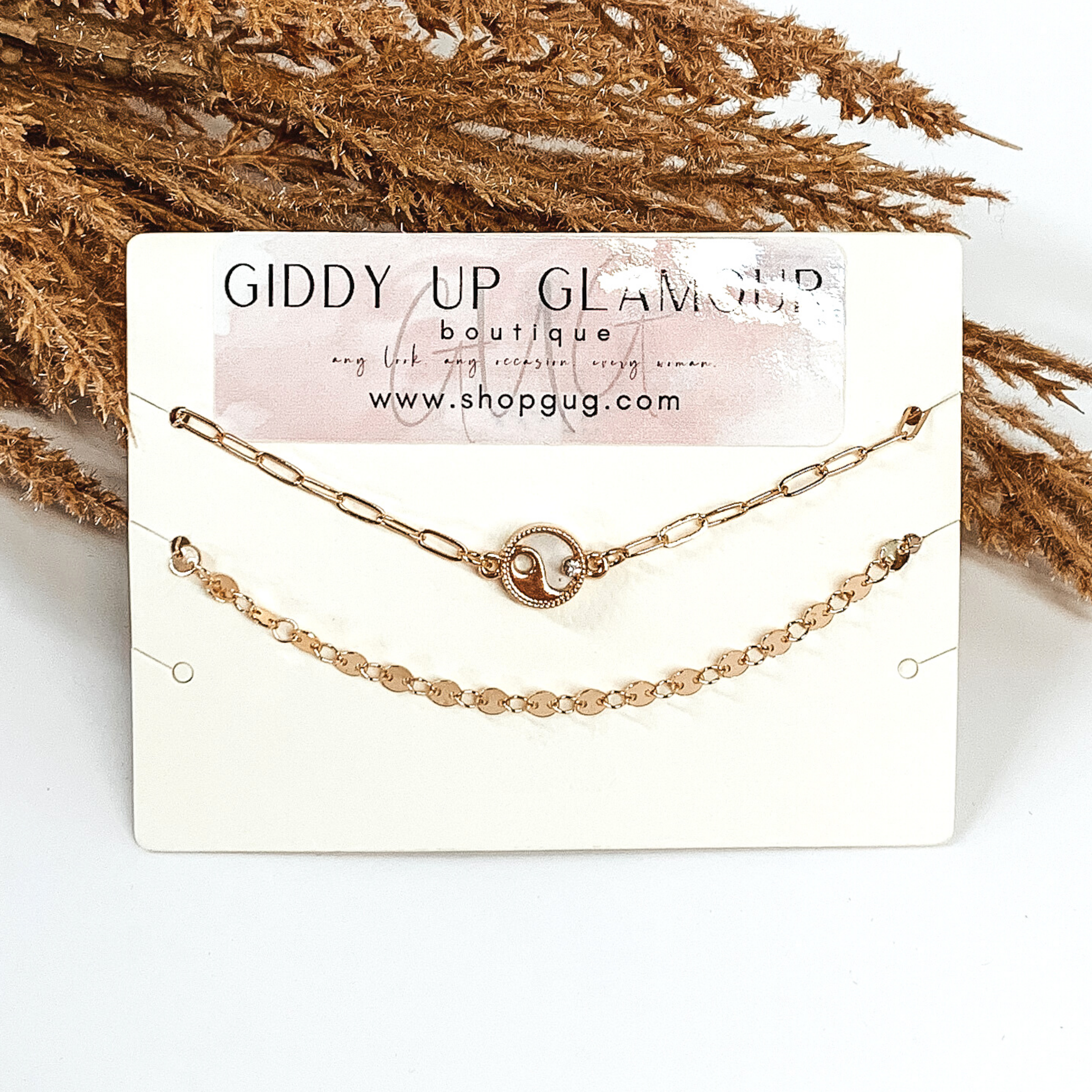 Two gold chained anklets. One is plain, while the second one has a yin-yang charm. They are pictured on a a white background that has some brown floral. 