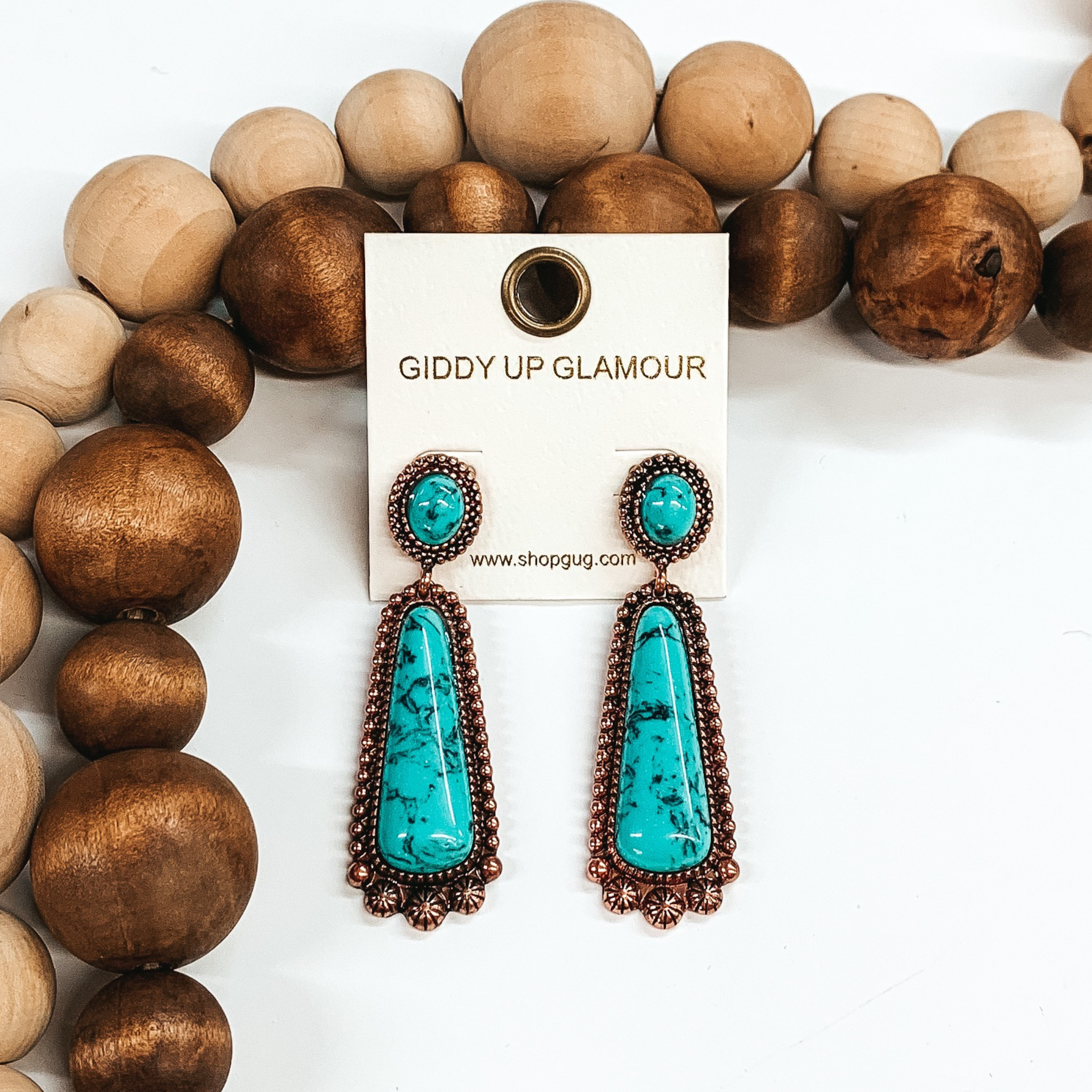 These earrings are copper and include an oval turquoise stud with a turquoise drop stone in a rounded triangle shape. These earrings are pictured on a white that has tan and brown beads on it.