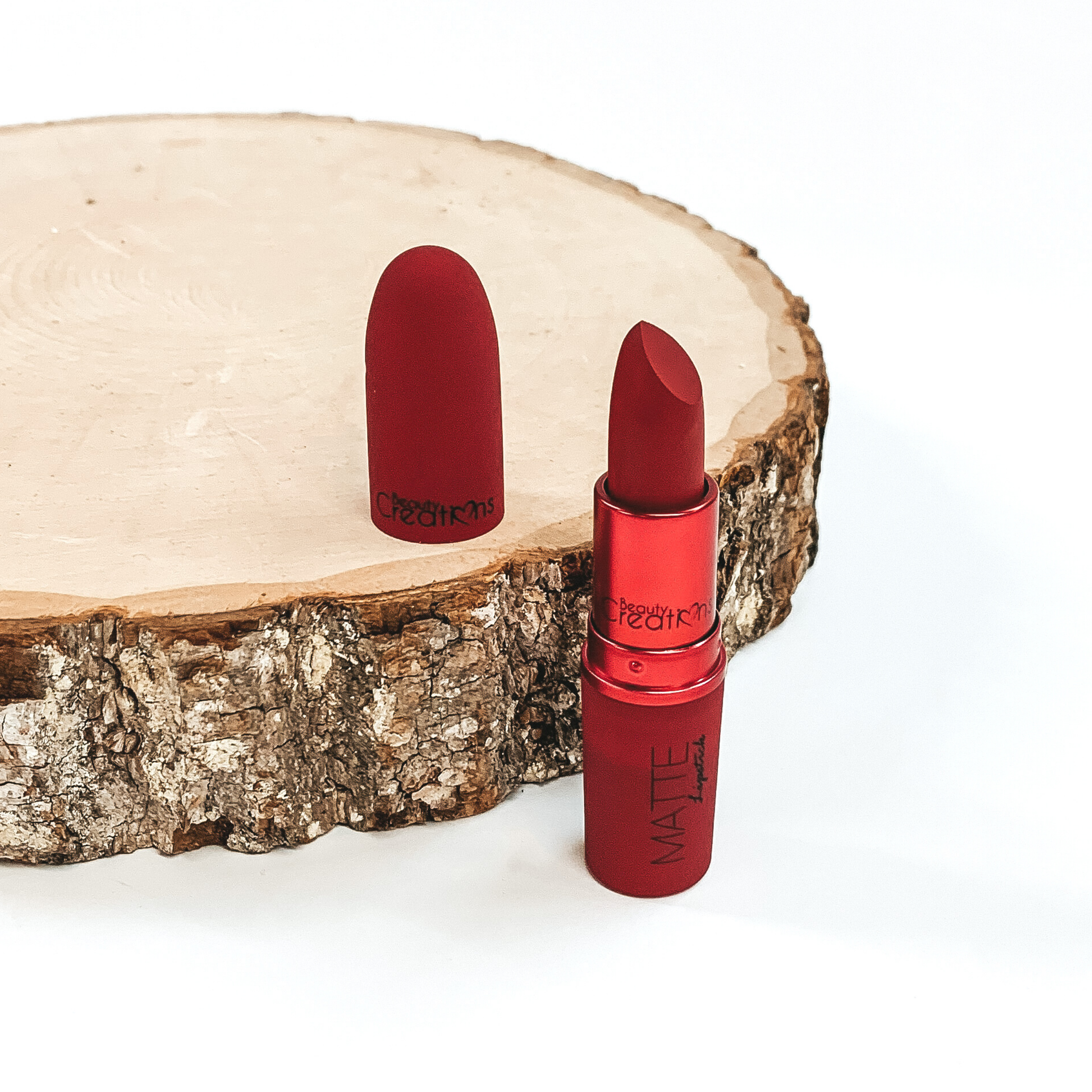 Shade my cherry lipstick that is opened and twisted up. The tube is standing on a white background and next to it is some wood that the lid from the lipstick is placed on.
