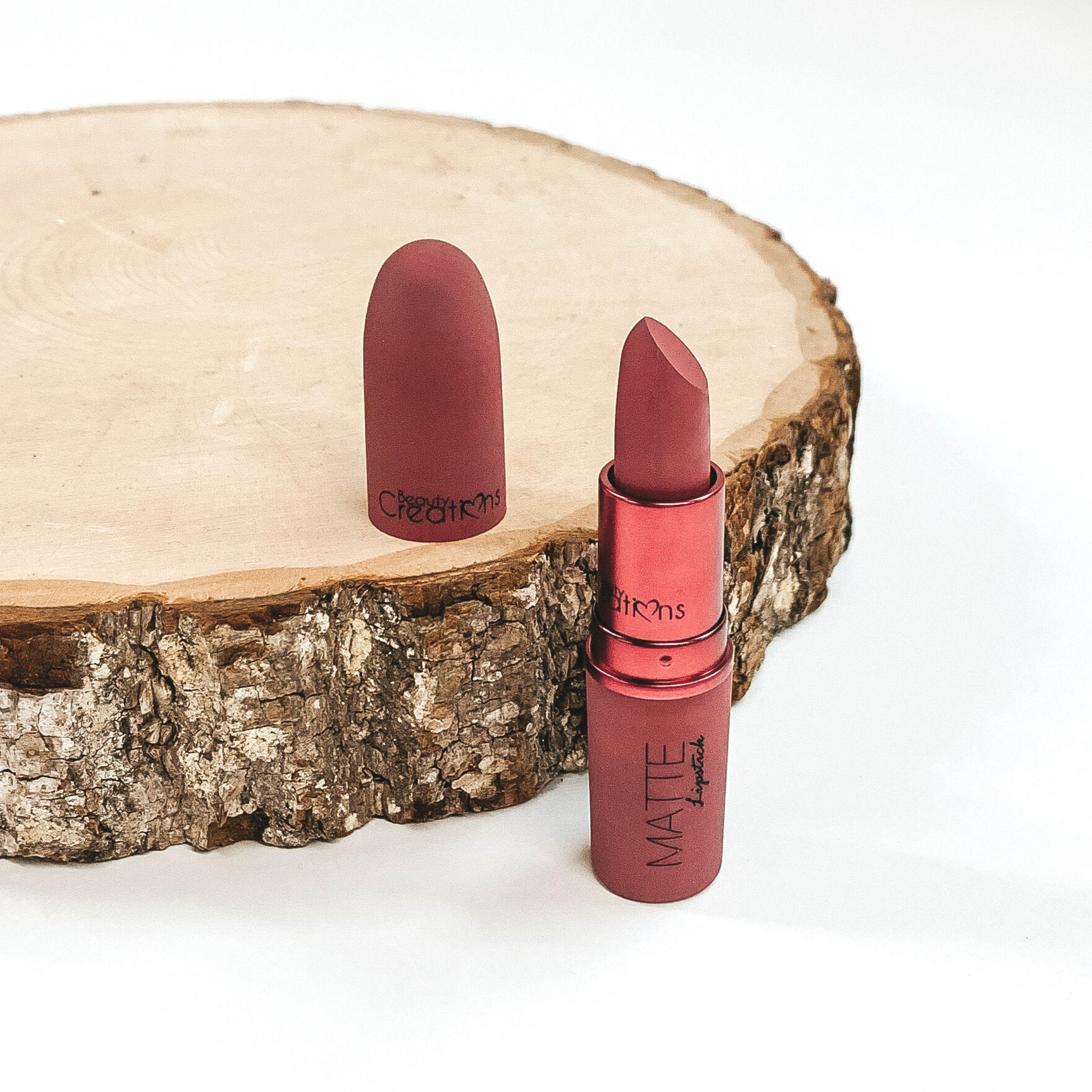 Shade kiss me lipstick that is opened and twisted up. The tube is standing on a white background and next to it is some wood that the lid from the lipstick is placed on.