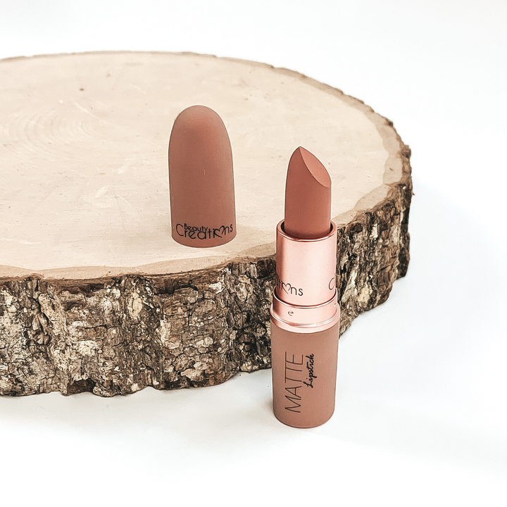 Shade obsessed lipstick that is opened and twisted up. The tube is standing on a white background and next to it is some wood that the lid from the lipstick is placed on.