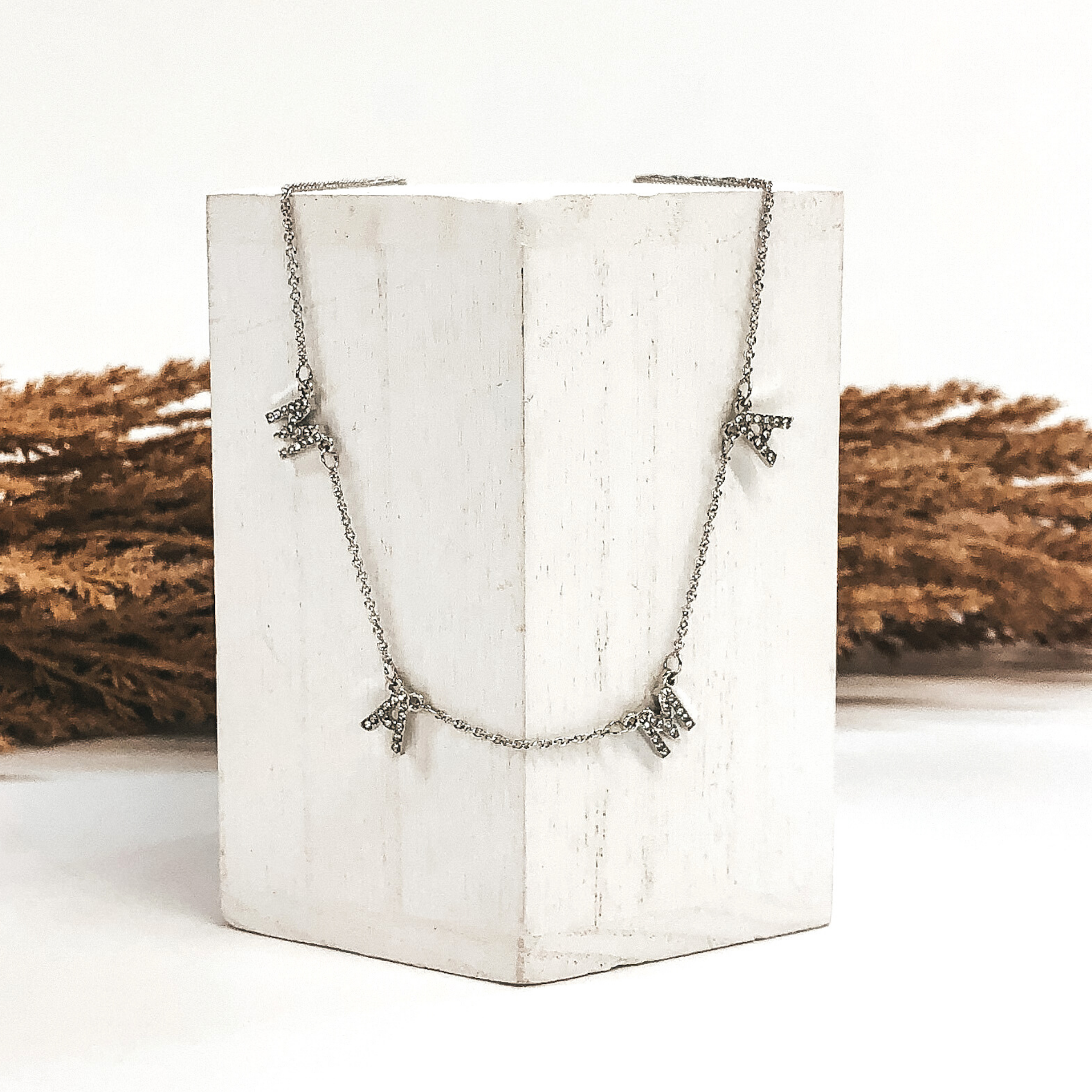 Silver necklace with crystal letter pendants to spell out "MAMA" laid on a white block in front of brown floral. 