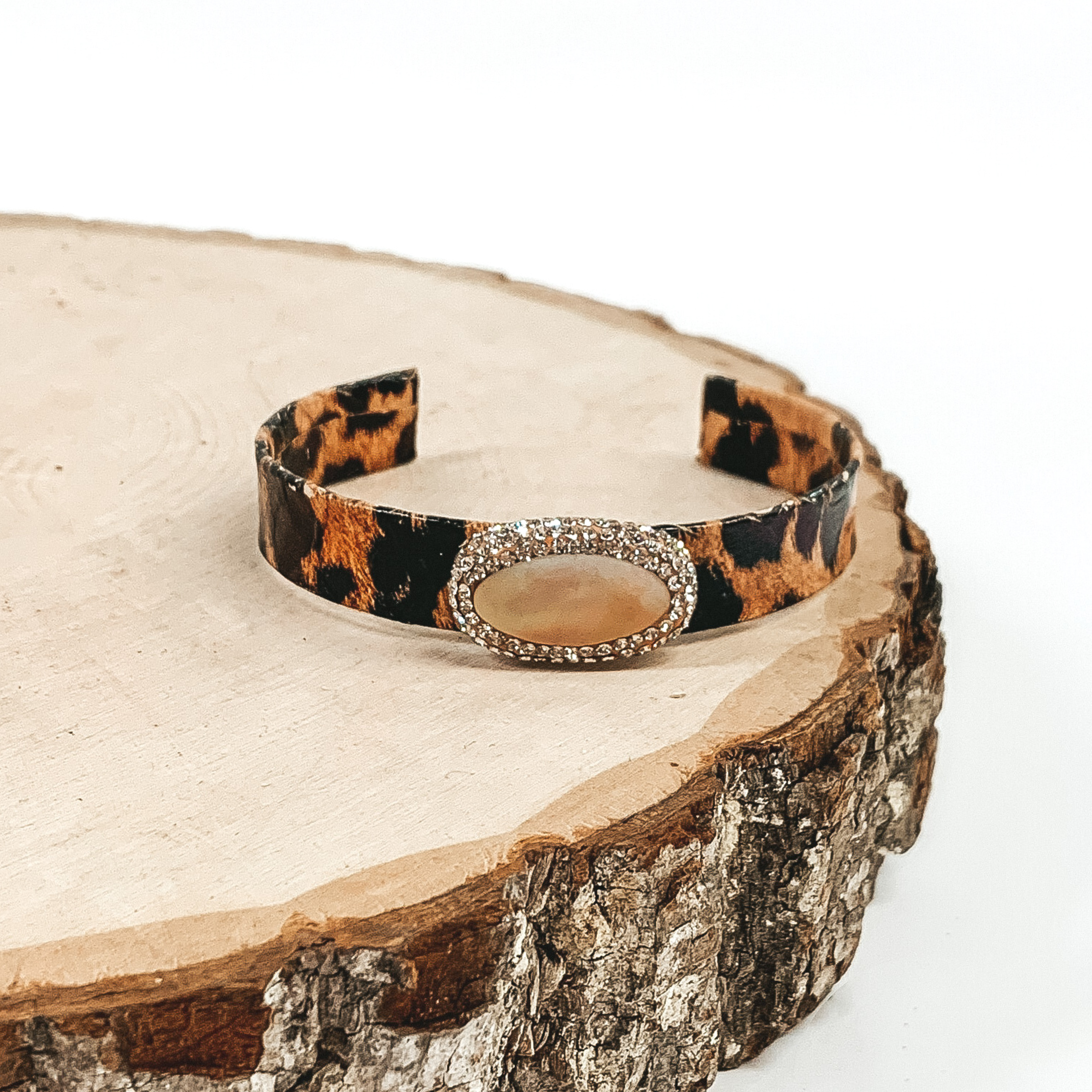 Leopard cuff with champagne oval stone that is outlined in crystals. This cuff is pictured on a piece of wood on a white background. 