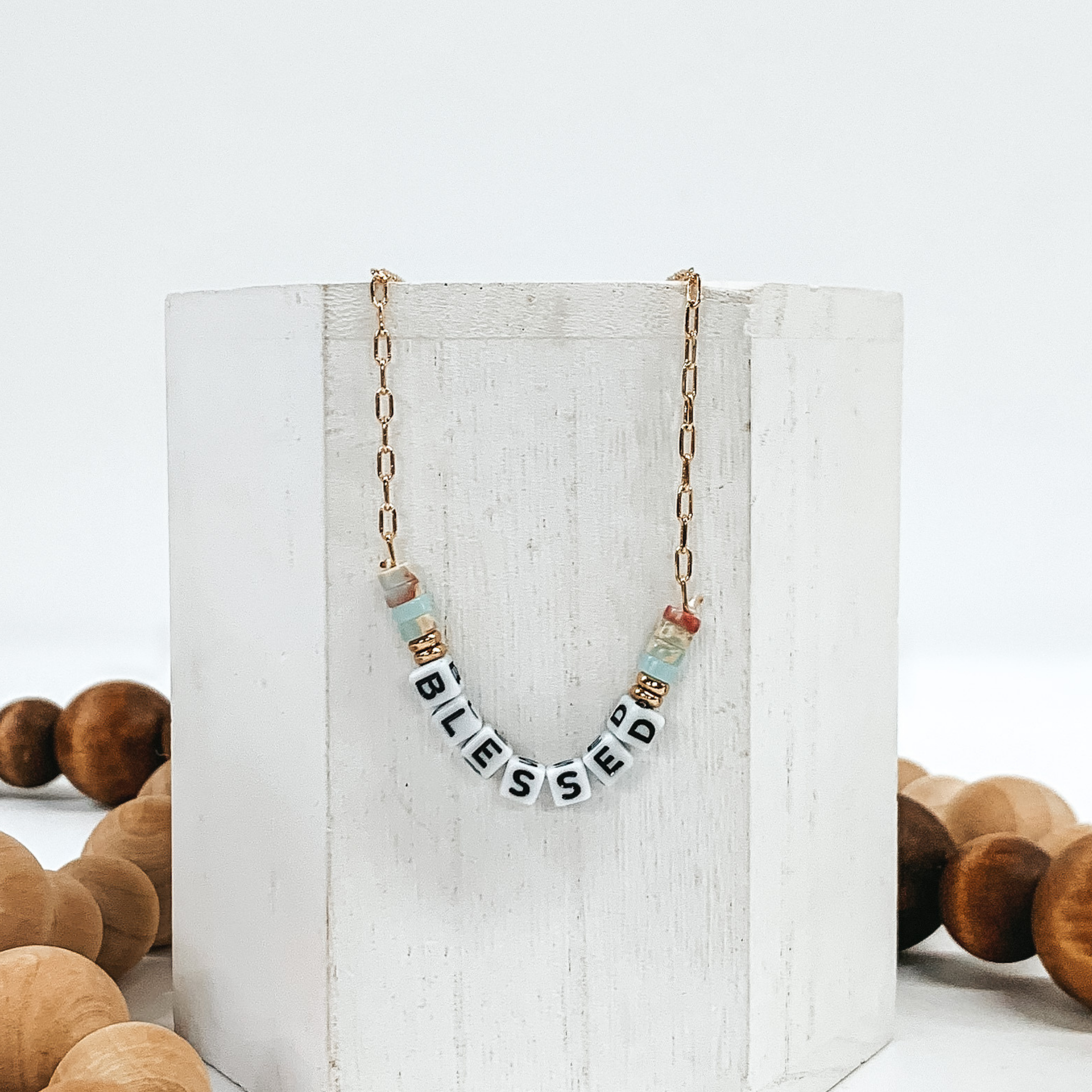 Small gold chain necklace with colorful marble beads, gold beads, and white letter beads that spell out "BLESSED". Pictured of a white block that is on a white background with tan and brown beads.