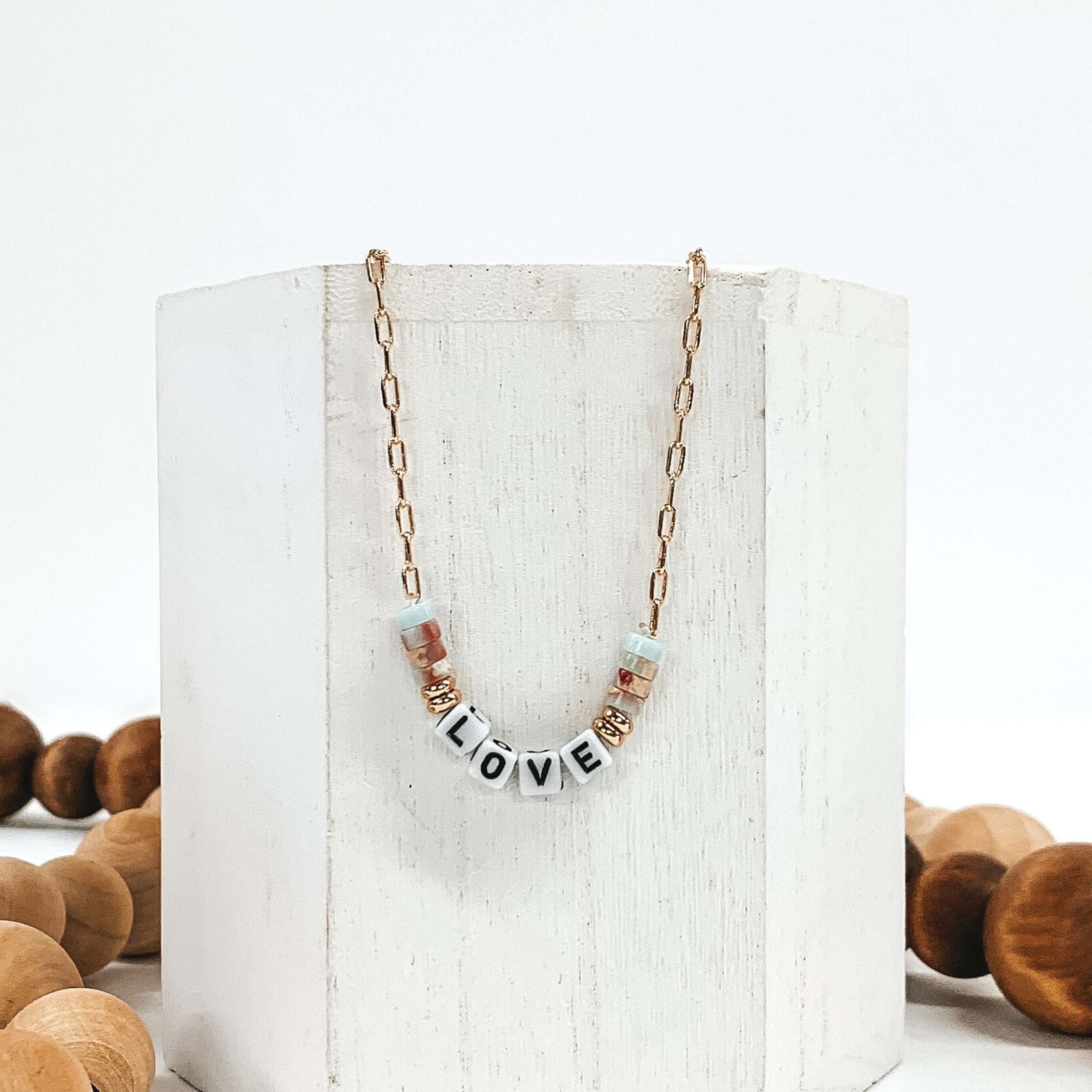 Small gold chain necklace with colorful marble beads, gold beads, and white letter beads that spell out "LOVE". Pictured of a white block that is on a white background with tan and brown beads.