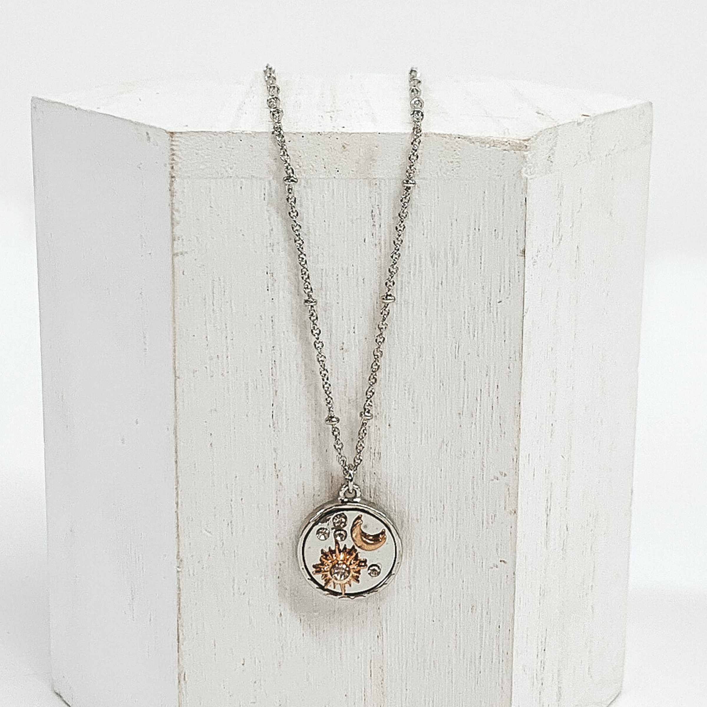 Silver necklace with silver circle pendant. The circle pendant has clear round circles, a gold star, and a gold moon. This necklace is pictured on a white block and on a white background. 