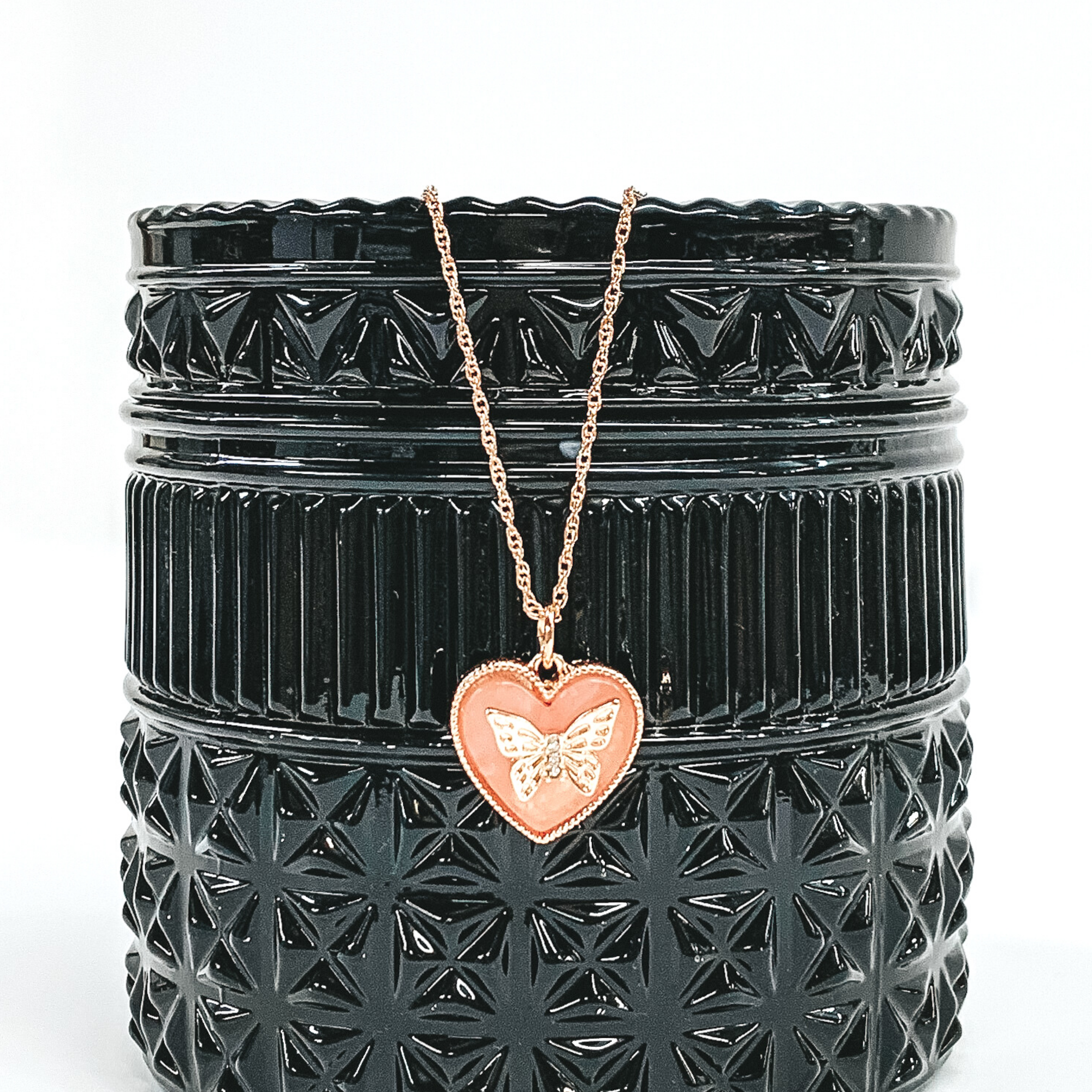 Gold necklace with blush colored heart pendant. The heart pendant has a gold butterfly charm in the center. The necklaces is pictured of a a black container that is in front of a white background. 