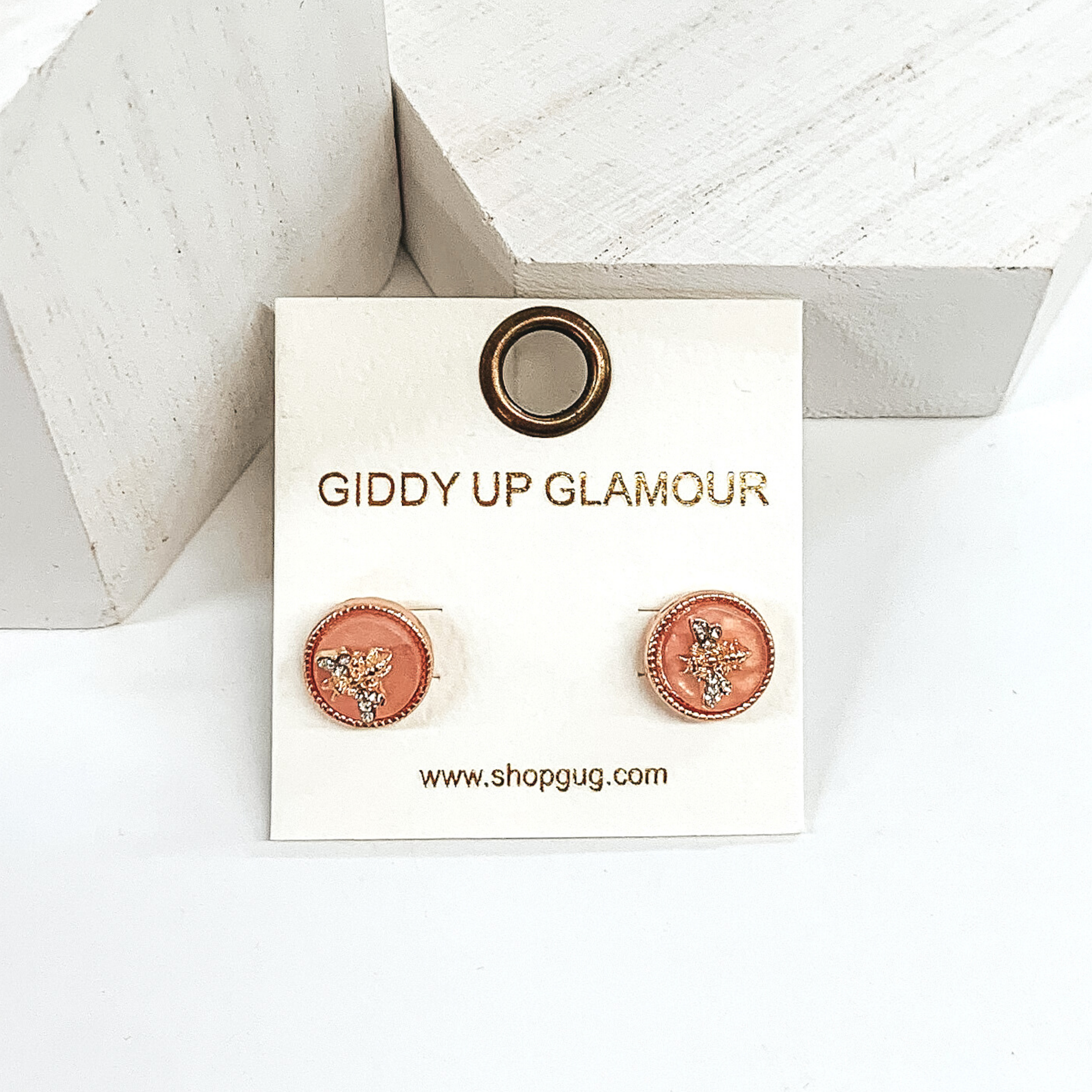 Blush circle stud earrings with a gold bee charm in the center. There are crystals on the wings of the bee charm. These studs are pictured of a white background.