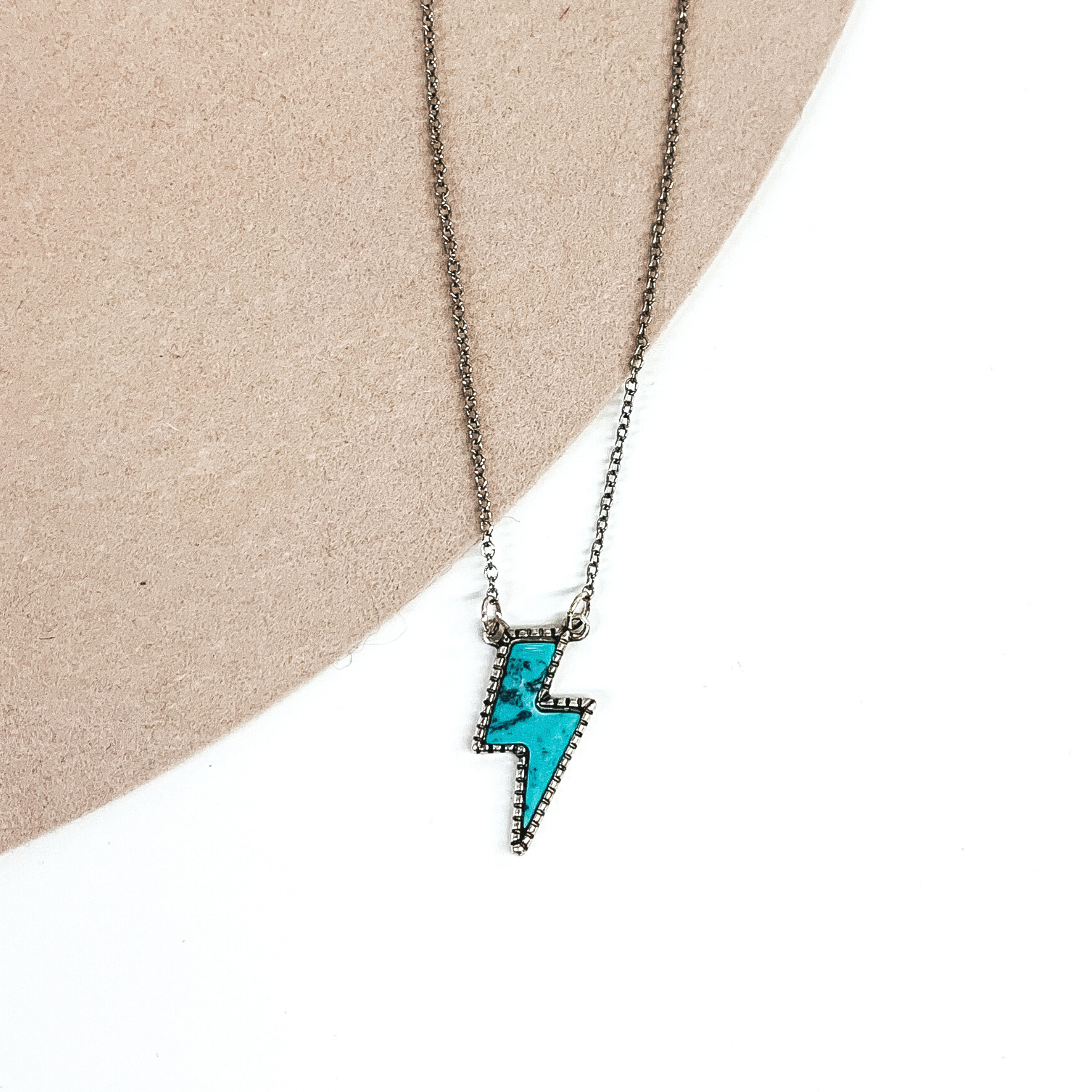 Silver necklace chain with turquoise lightning bolt shaped pendant. This necklace is pictured on a white and beige background. 