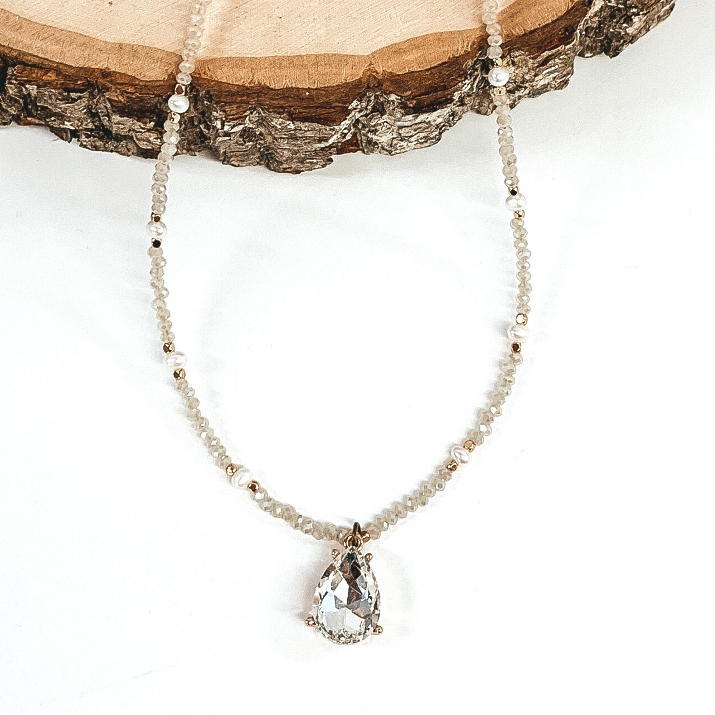 Clear crystal beads, white pearls, and gold beaded necklace with a clear teardrop crystal. This necklace is pictured on a white background with  wood at the top.