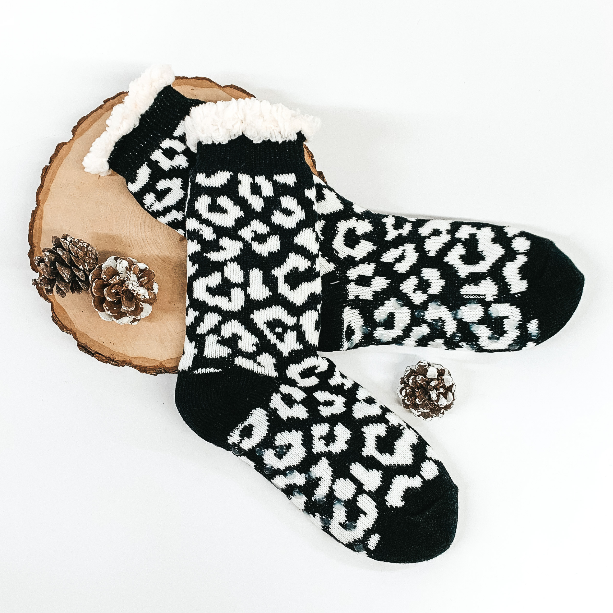 Black socks with white leopard spots. The socks has white sherpa at the top of the socks. The socks are pictured on a white background laying on a piece of wood with a few pine cones.