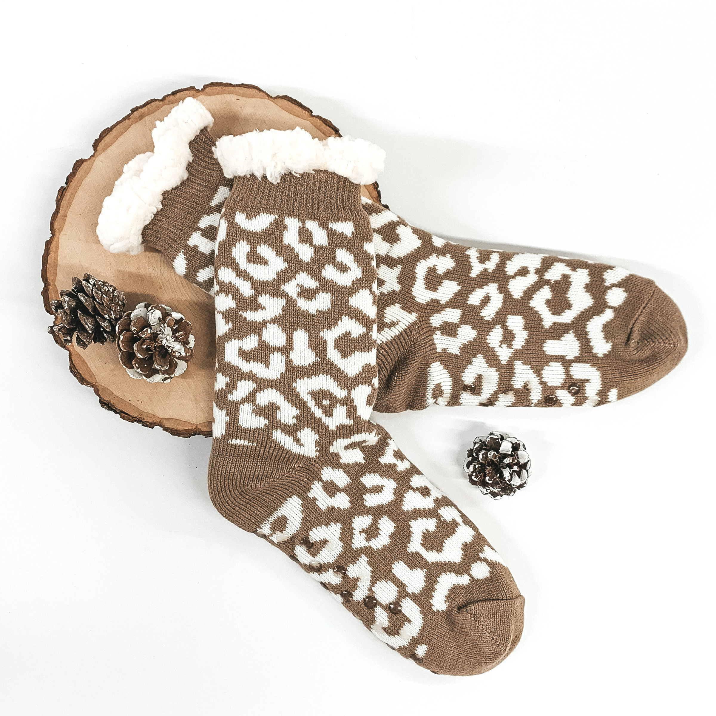 Beige socks with white leopard spots. The socks has white sherpa at the top of the socks. The socks are pictured on a white background laying on a piece of wood with a few pine cones. 