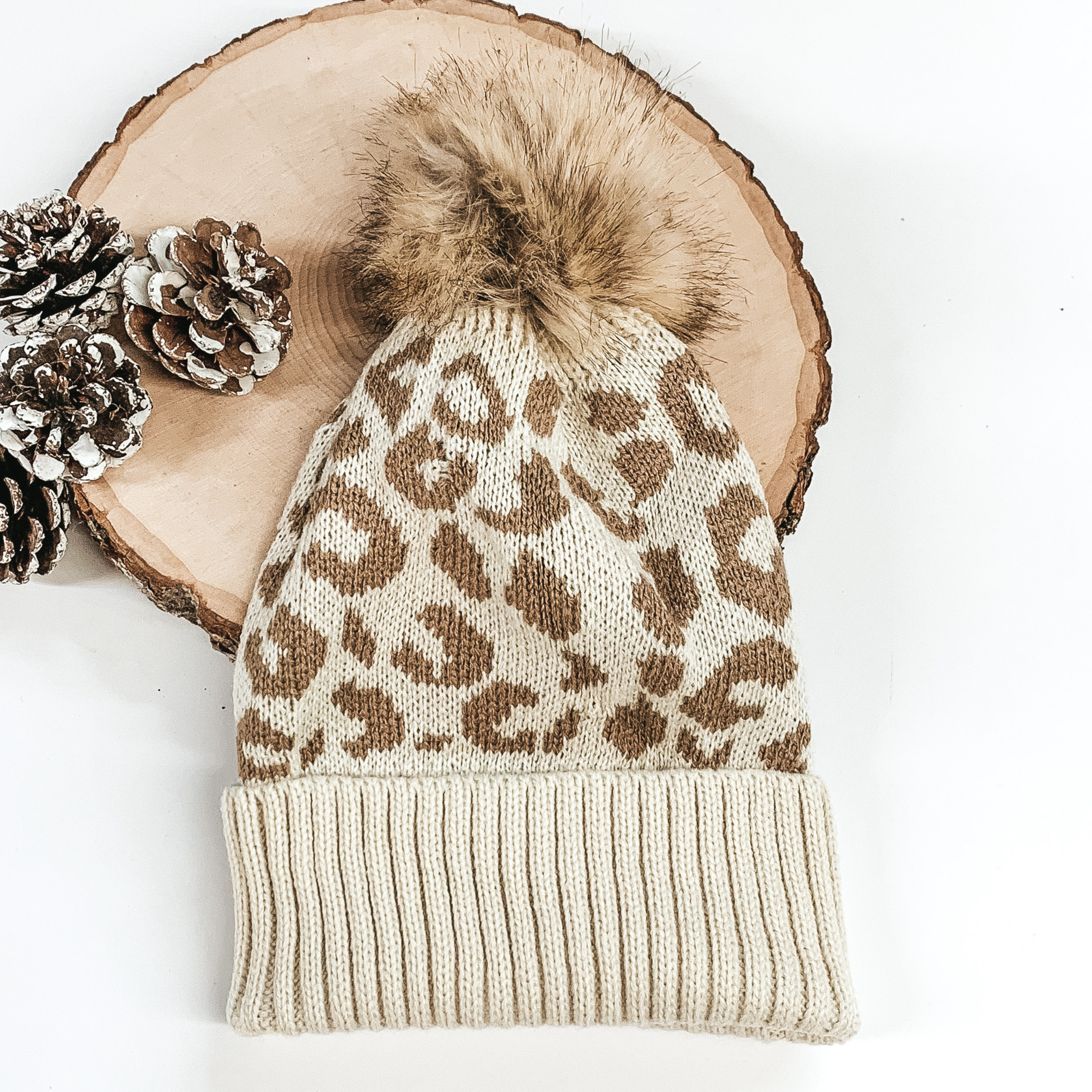 Ivory beanie with tan leopard print and tan pom pom at the top. This beanie is pictured laying on a piece of wood on a white background. 