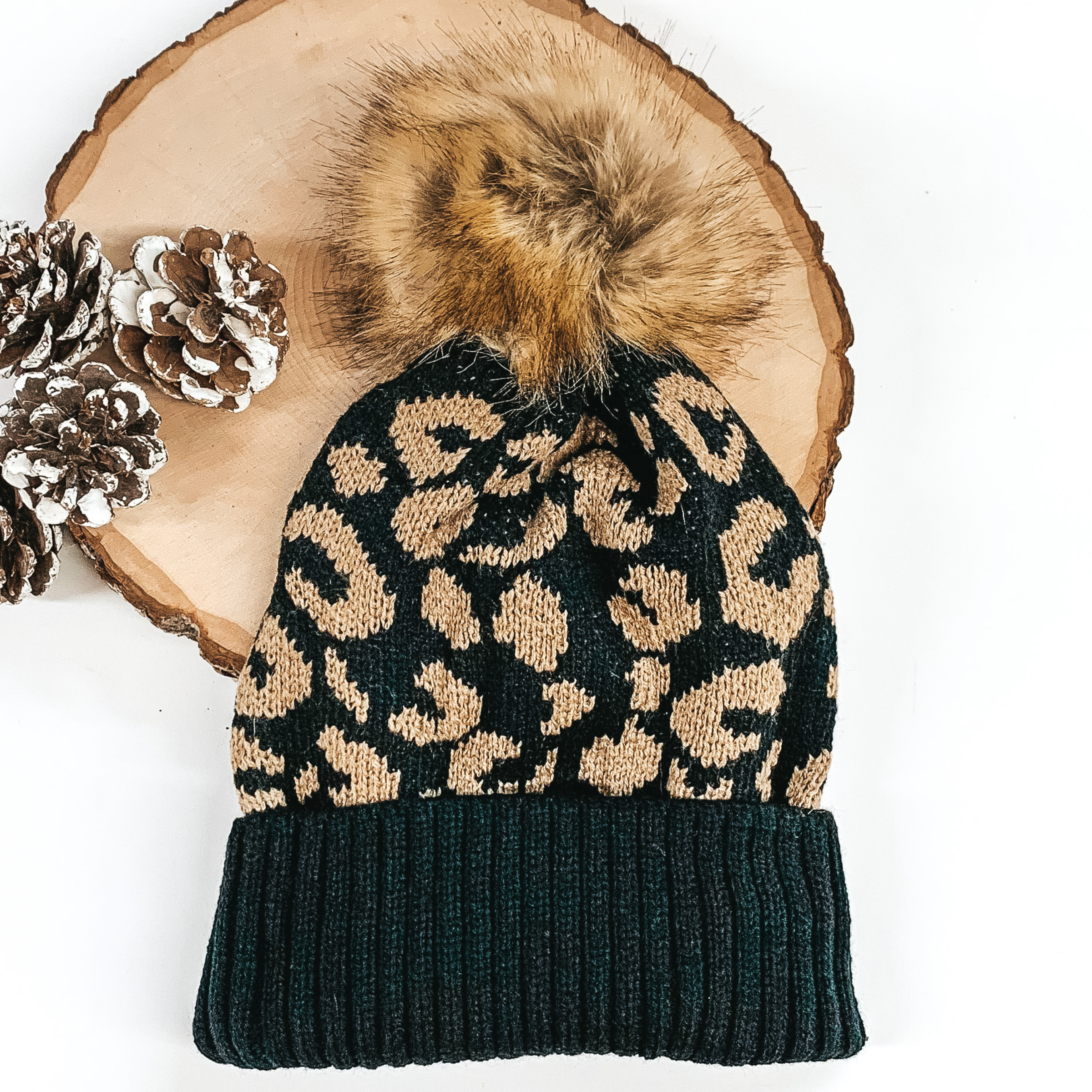 Black beanie with tan leopard print and tan pom pom at the top. This beanie is pictured laying on a piece of wood on a white background.