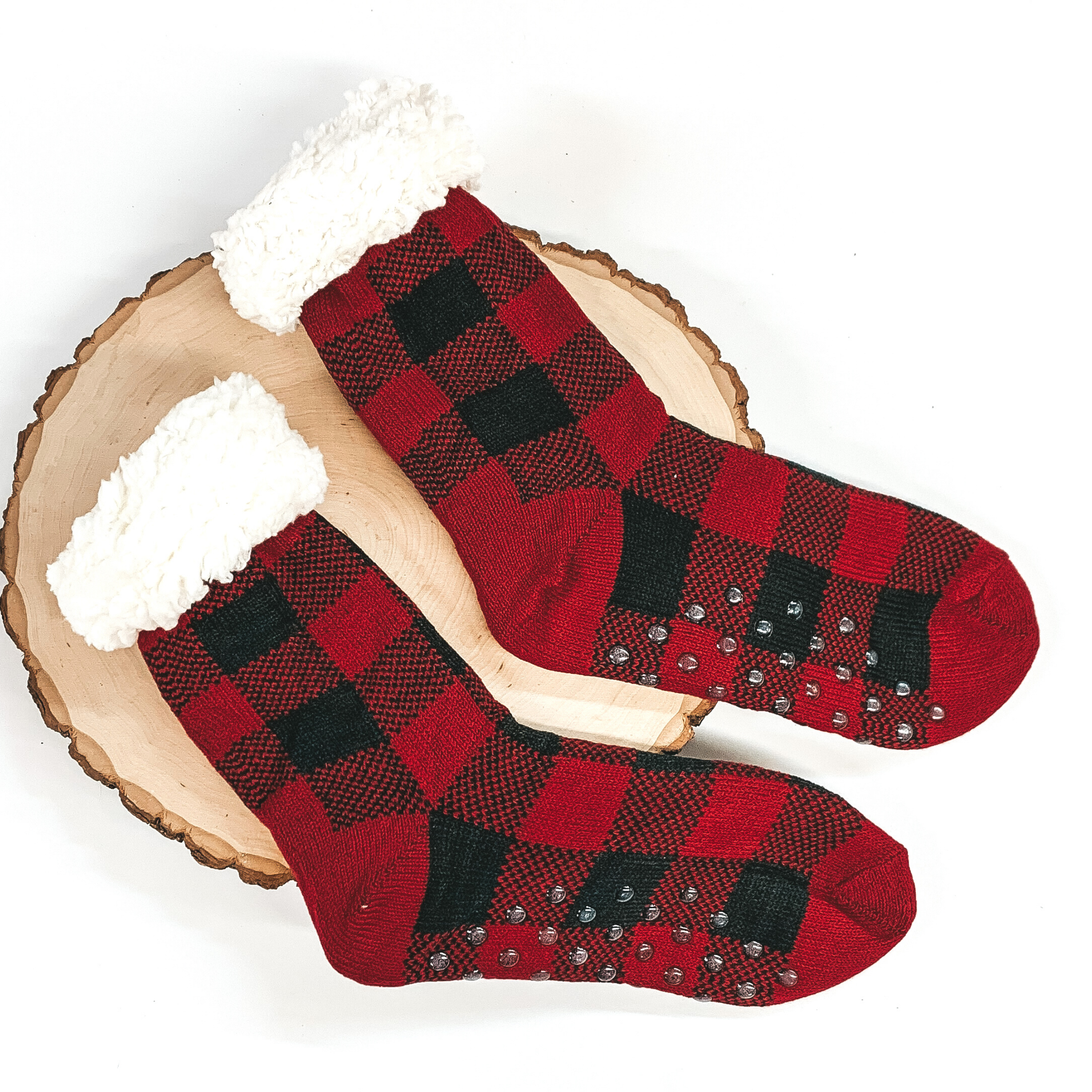Red buffalo plaid socks that has rubber grips on the bottom and white fluff on the top of the socks. these socks are pictured laying on a piece of wood on a white background. 