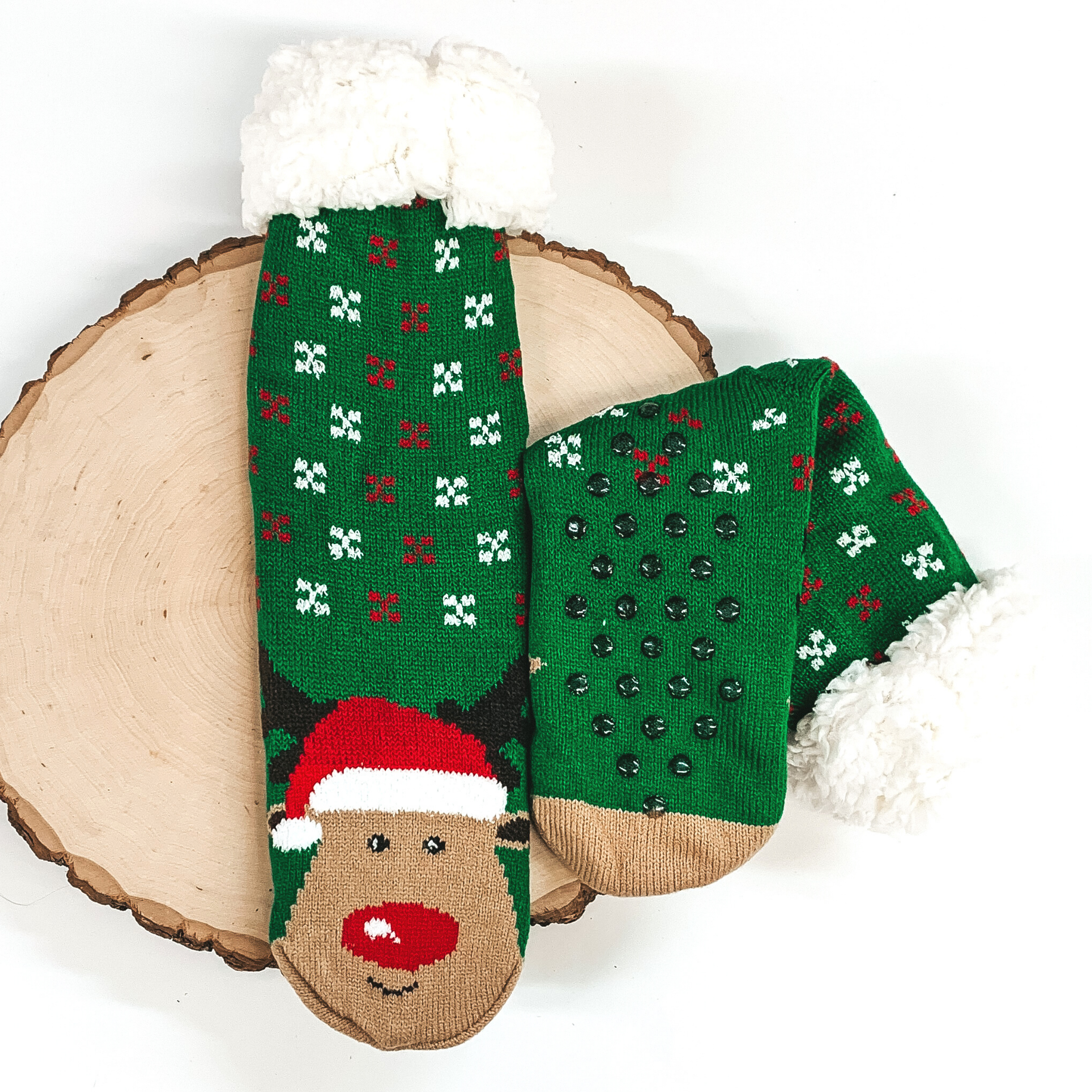 Green socks that has Rudolph's face with a santa hat towards the bottom of the socks. These socks also have red and white snow flakes on the ankle parts, has rubber grips on the bottom, and white fluff on the top of the socks. these socks are pictured laying on a piece of wood on a white background.
