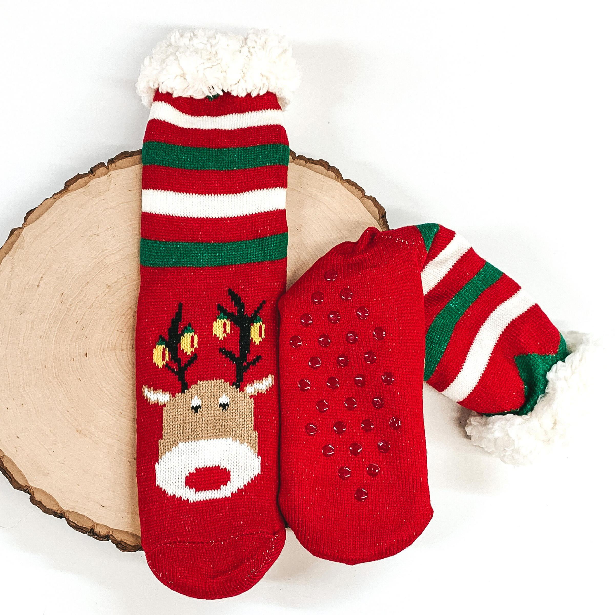 Red socks that has Rudolph's face with lights on the antlers towards the bottom of the socks. These socks also have green and white stripes on the ankle parts, has rubber grips on the bottom, and white fluff on the top of the socks. these socks are pictured laying on a piece of wood on a white background.