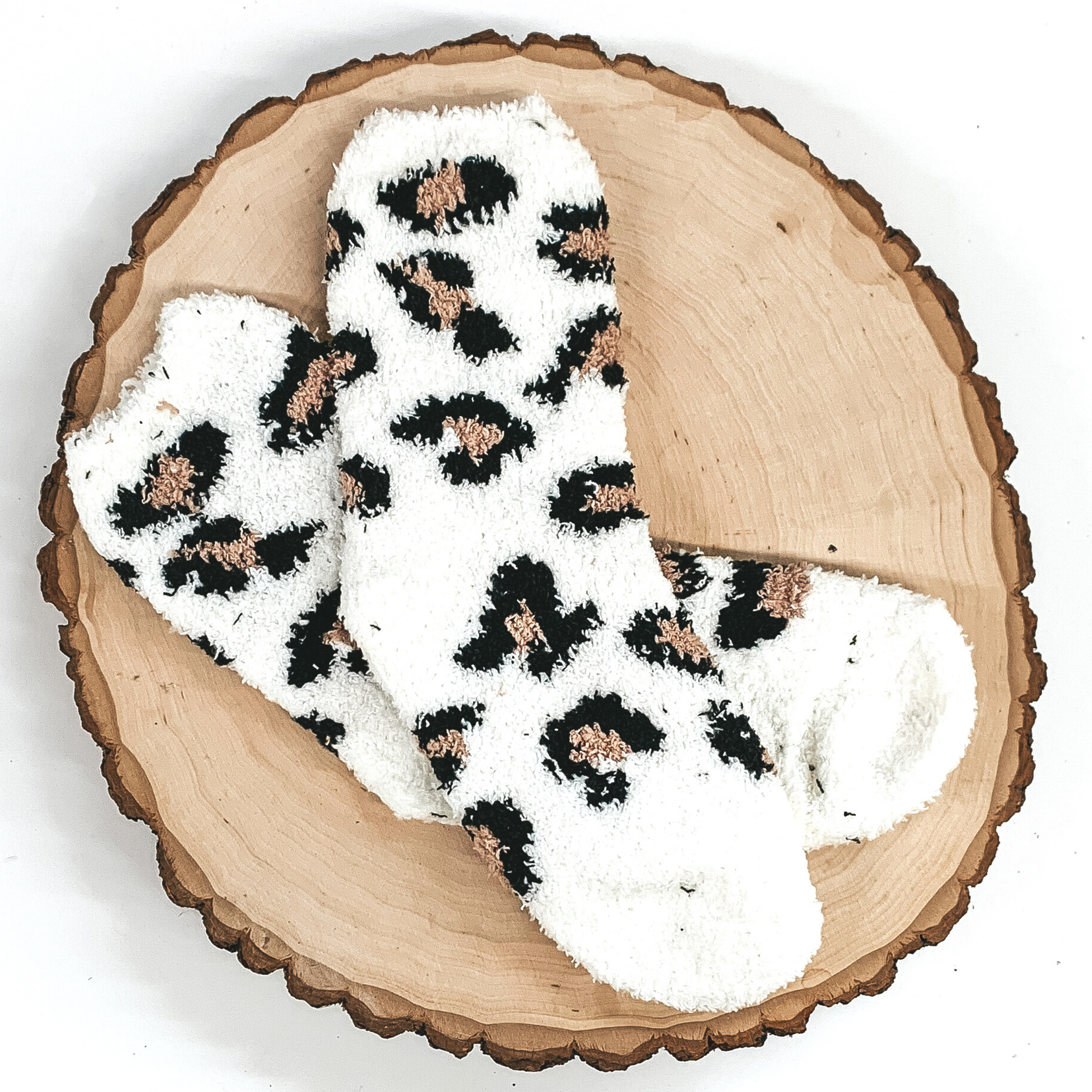 Fuzzy white socks with black and tan leopard print. These socks are pictured laying on a piece of wood that is on a white background.