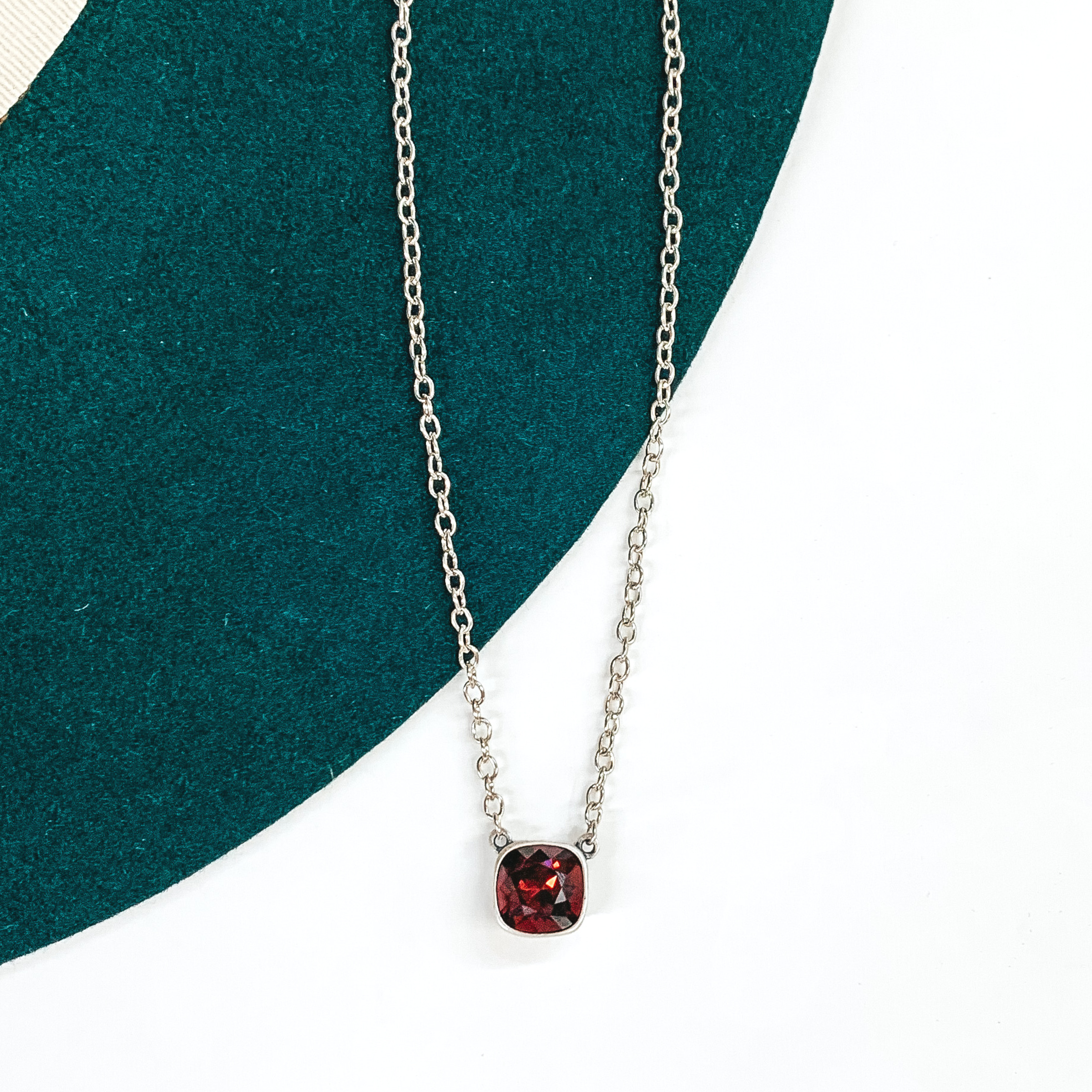 Silver chained necklace with a single maroon crystal. This necklace is pictured on a white and green background.
