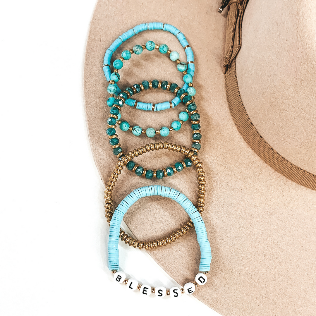 This bracelet set has a mix of turquoise disk beaded bracelets, turquoise circle beaded bracelet, a gold beaded bracelet, and a turquoise crystal beaded bracelet. One of the turquoise disk beaded bracelet has letter beads that spell out "BLESSED." These bracelets are pictured on a white and beige background.