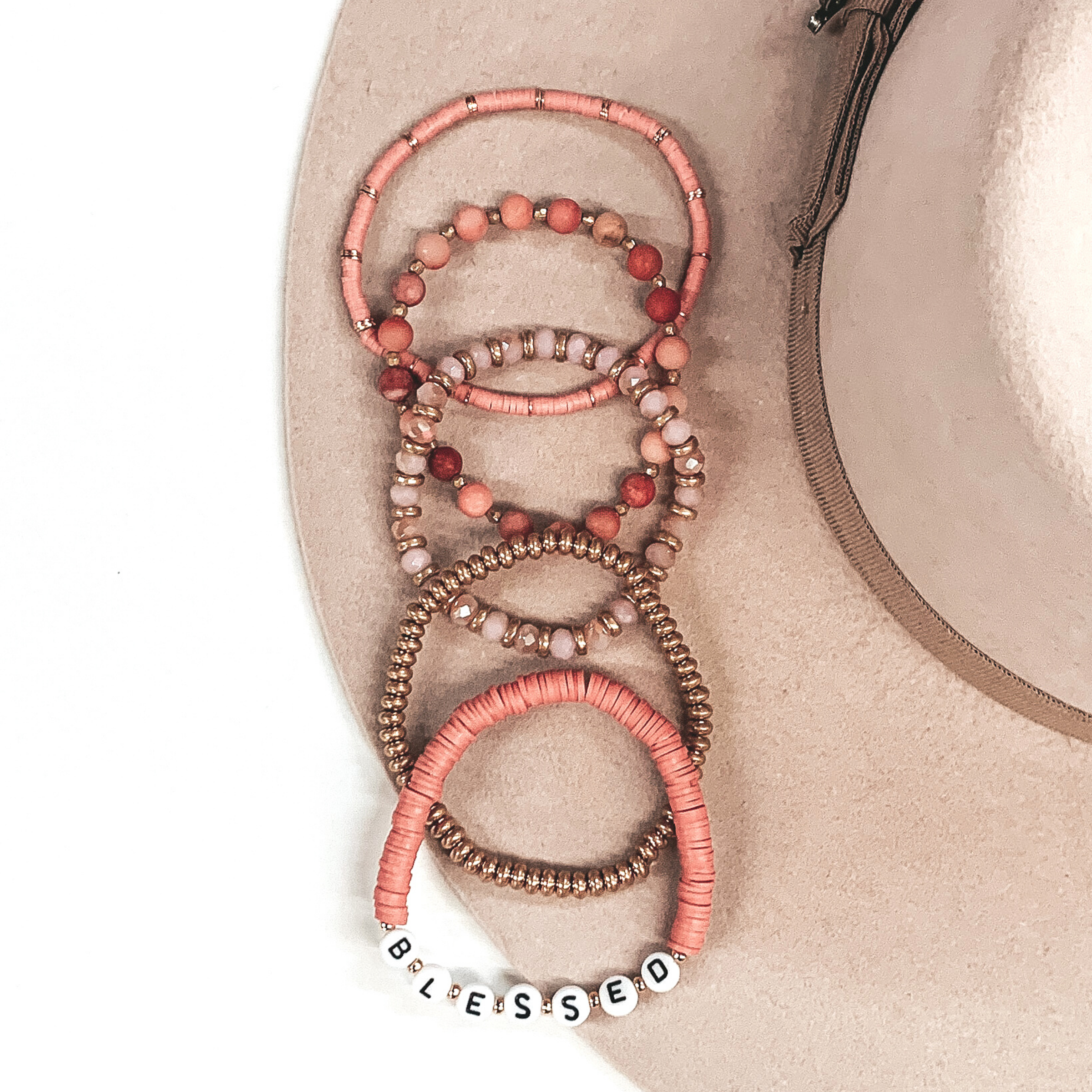 This bracelet set has a mix of blush disk beaded bracelets, blush circle beaded bracelet, a gold beaded bracelet, and a blush crystal beaded bracelet. One of the blush disk beaded bracelet has letter beads that spell out "BLESSED." These bracelets are pictured on a white and beige background.