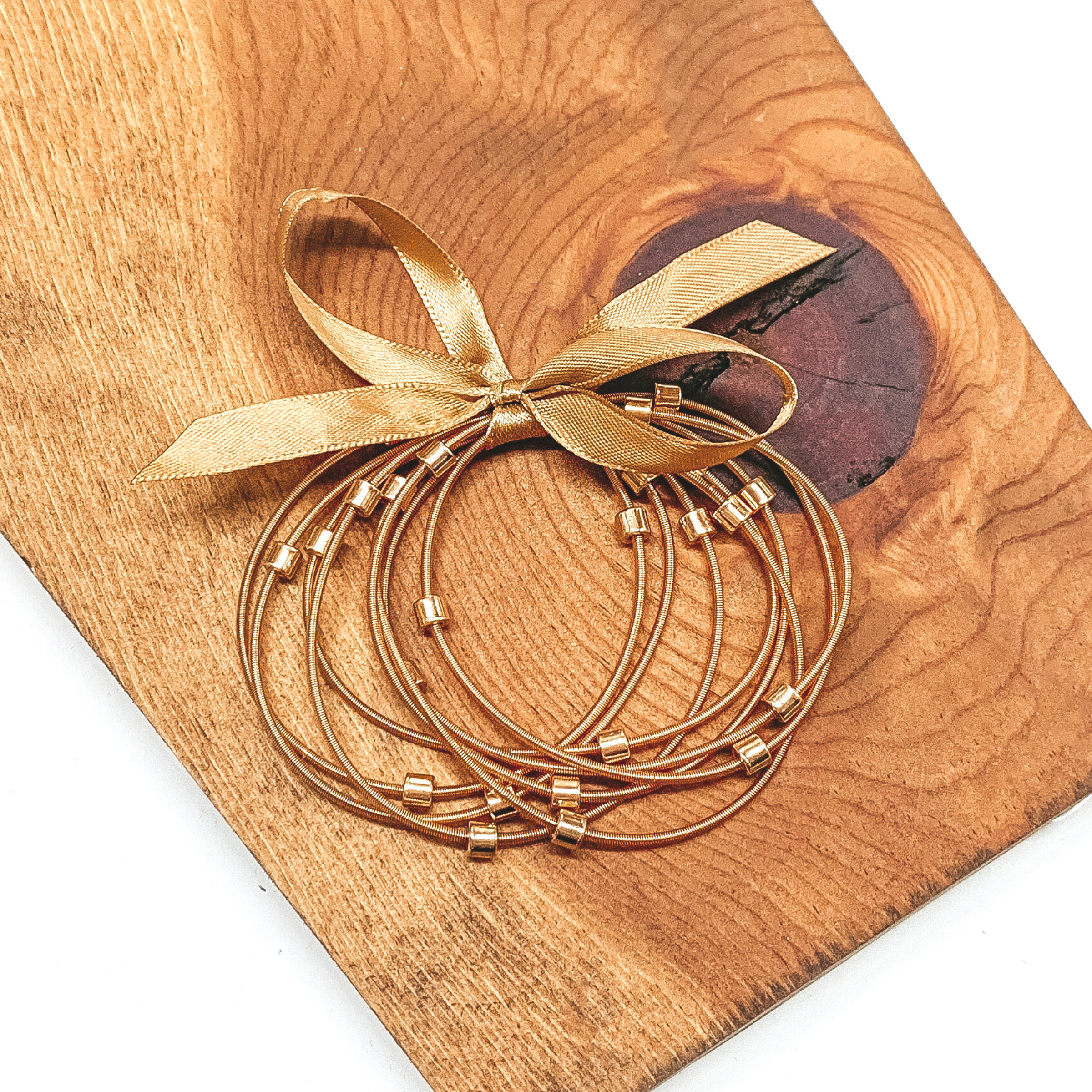 Group of gold spring wire elastic bracelet set tied together with a gold ribbon that is tied in a bow. All of the bracelets have small gold beads. This bracelet set is pictured laying next on a piece of wood on a white background.