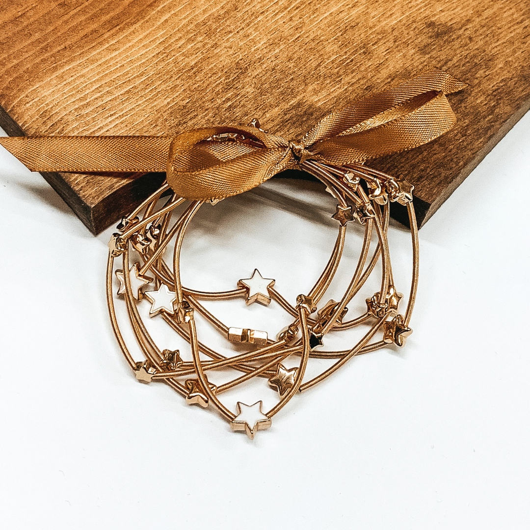 Group of gold spring wire elastic bracelet set tied together with a gold ribbon that is tied in a bow. All of the bracelets have small gold star beads. This bracelet set is pictured laying next on a piece of wood on a white background.