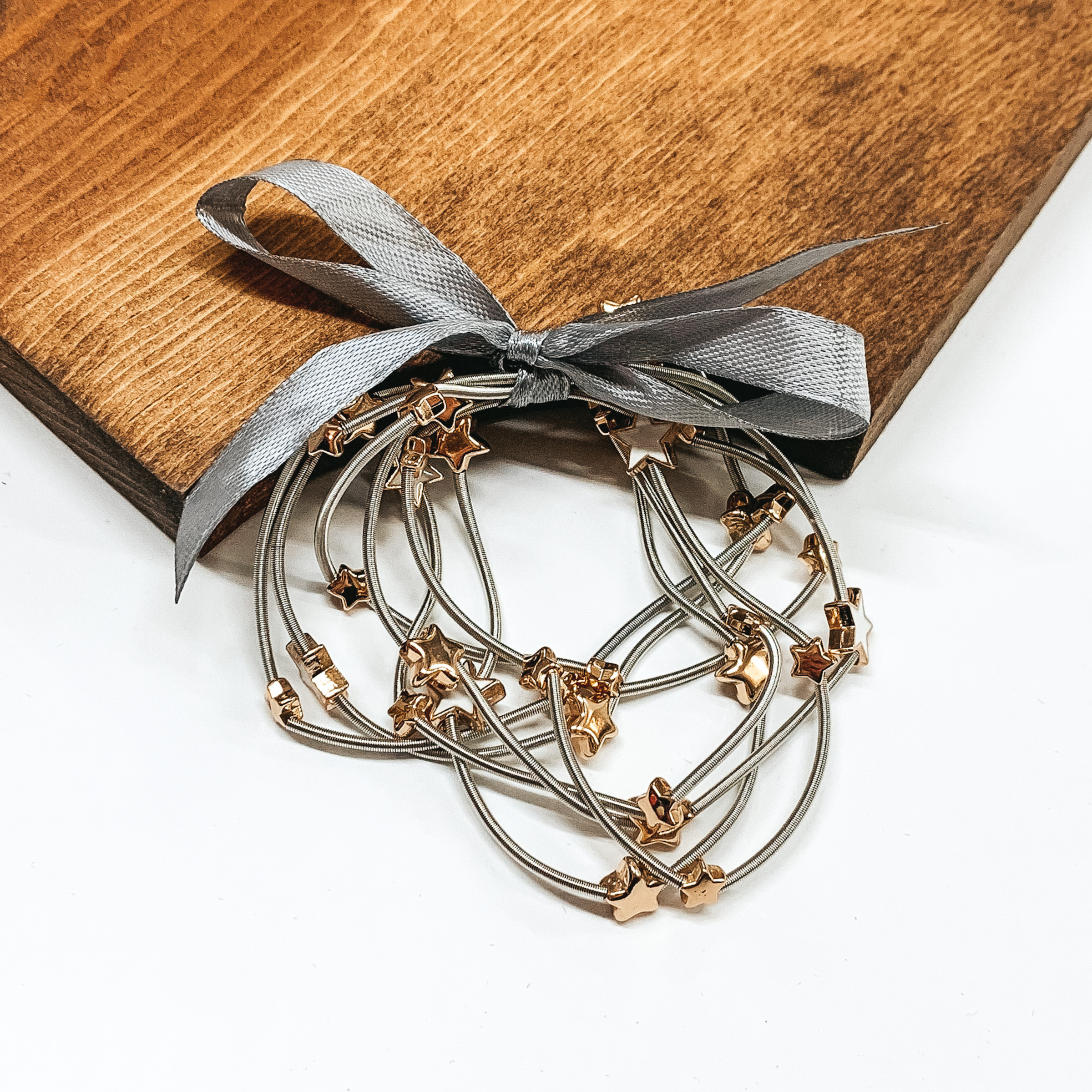Group of silver spring wire elastic bracelet set tied together with a silver ribbon that is tied in a bow. All of the bracelets have small gold star beads. This bracelet set is pictured laying next on a piece of wood on a white background.