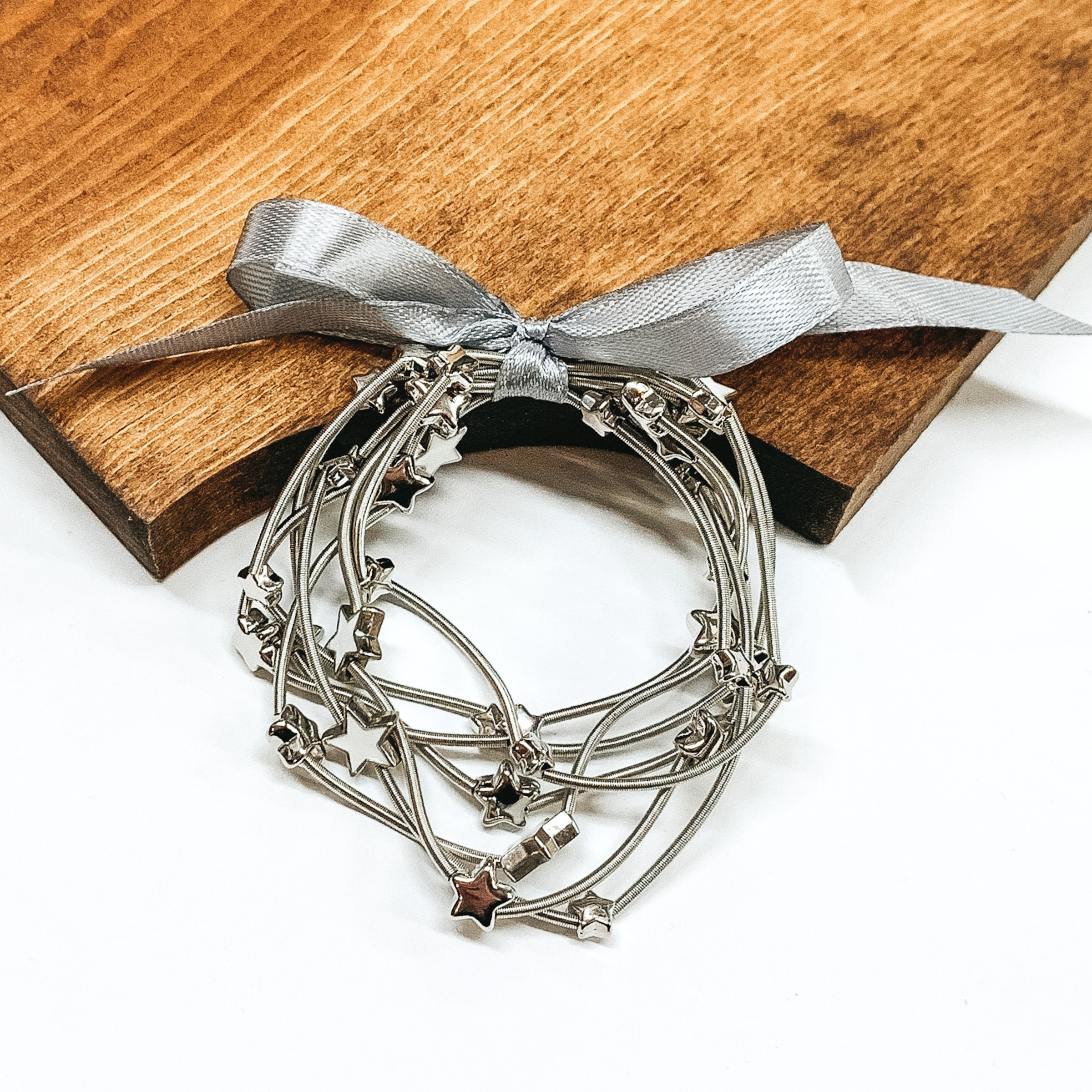 Group of silver spring wire elastic bracelet set tied together with a silver ribbon that is tied in a bow. All of the bracelets have small silver star beads. This bracelet set is pictured laying next on a piece of wood on a white background.