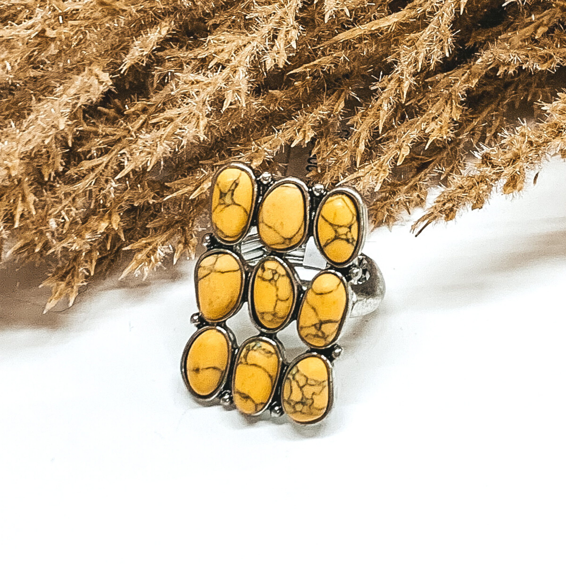This is a silver ring with nine yellow stones in rows of three creating a rectangle shape. This ring is pictured on a white background with some tan floral in the background.