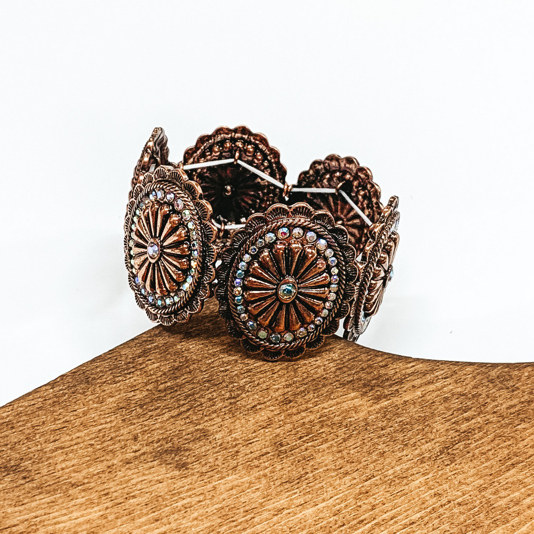 Copper colored oval concho bracelet with a small crystal center stone and a crystal outline. This bracelet is pictured laying on a piece of wood on a white background.