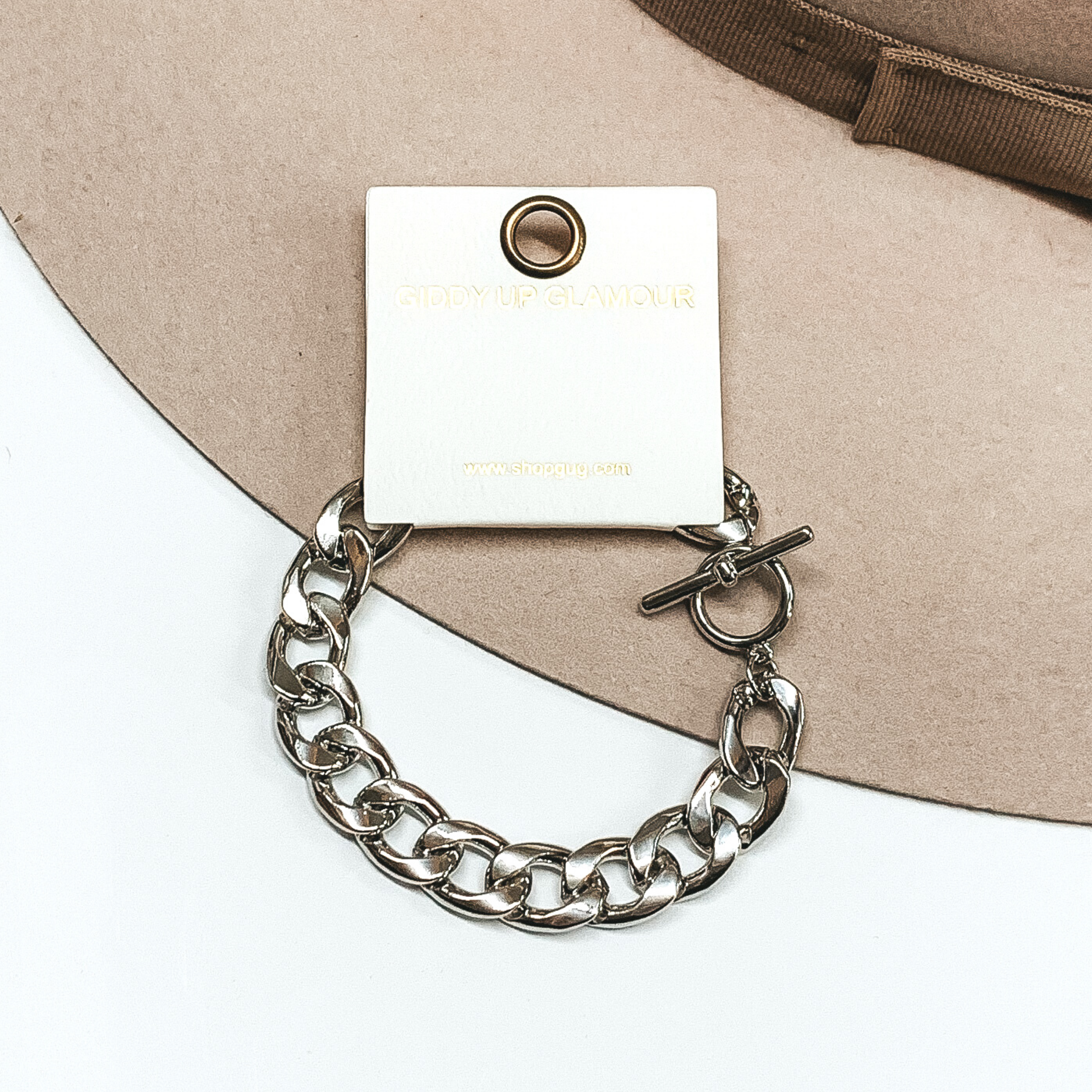 This silver chunky chain bracelet has a bar toggle clasp and is on a square tag. This bracelet is pictured on a white and beige background. 