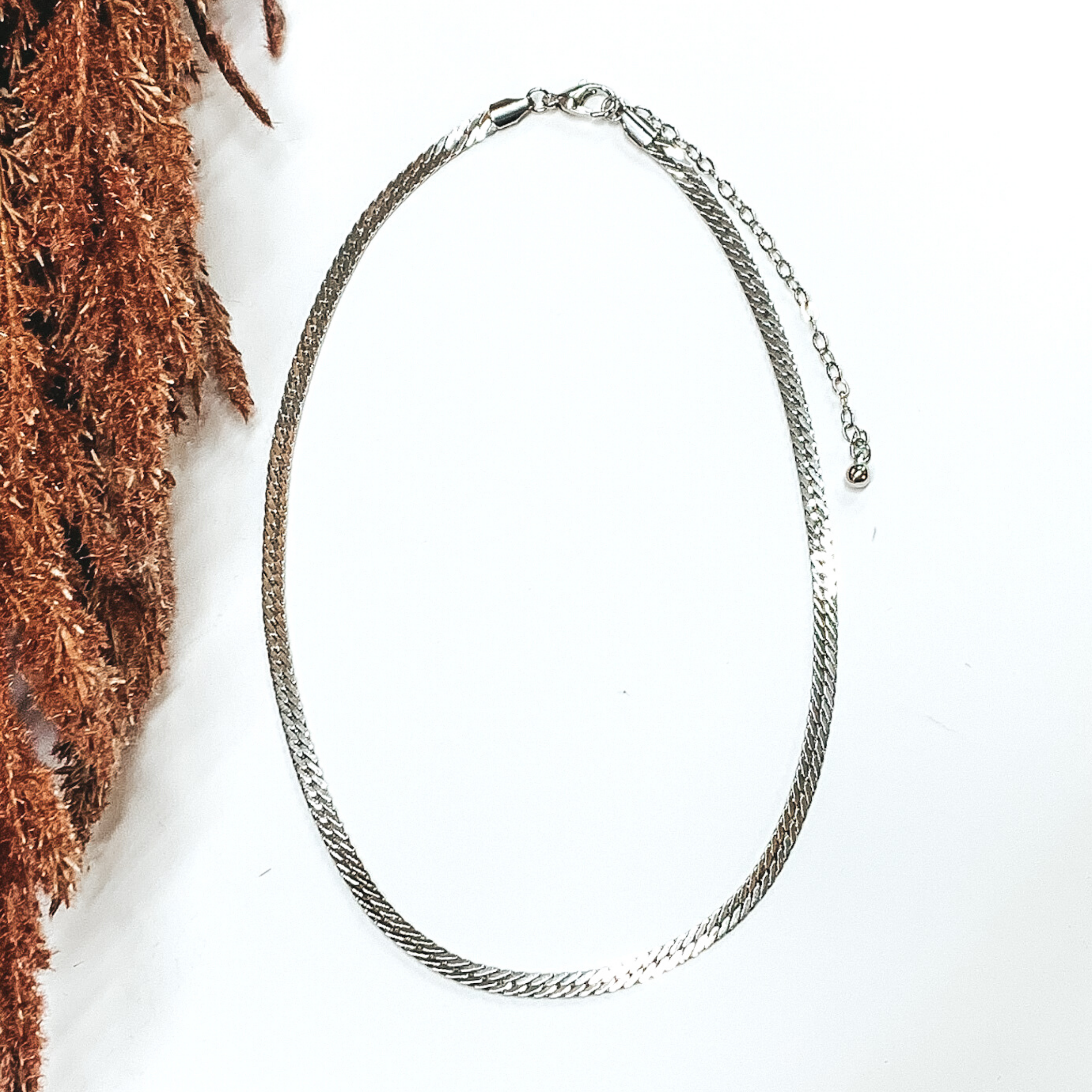 Simple flat snake chain necklace in silver. This necklace is pictured on a white background with brown floral on the side of the pic. 