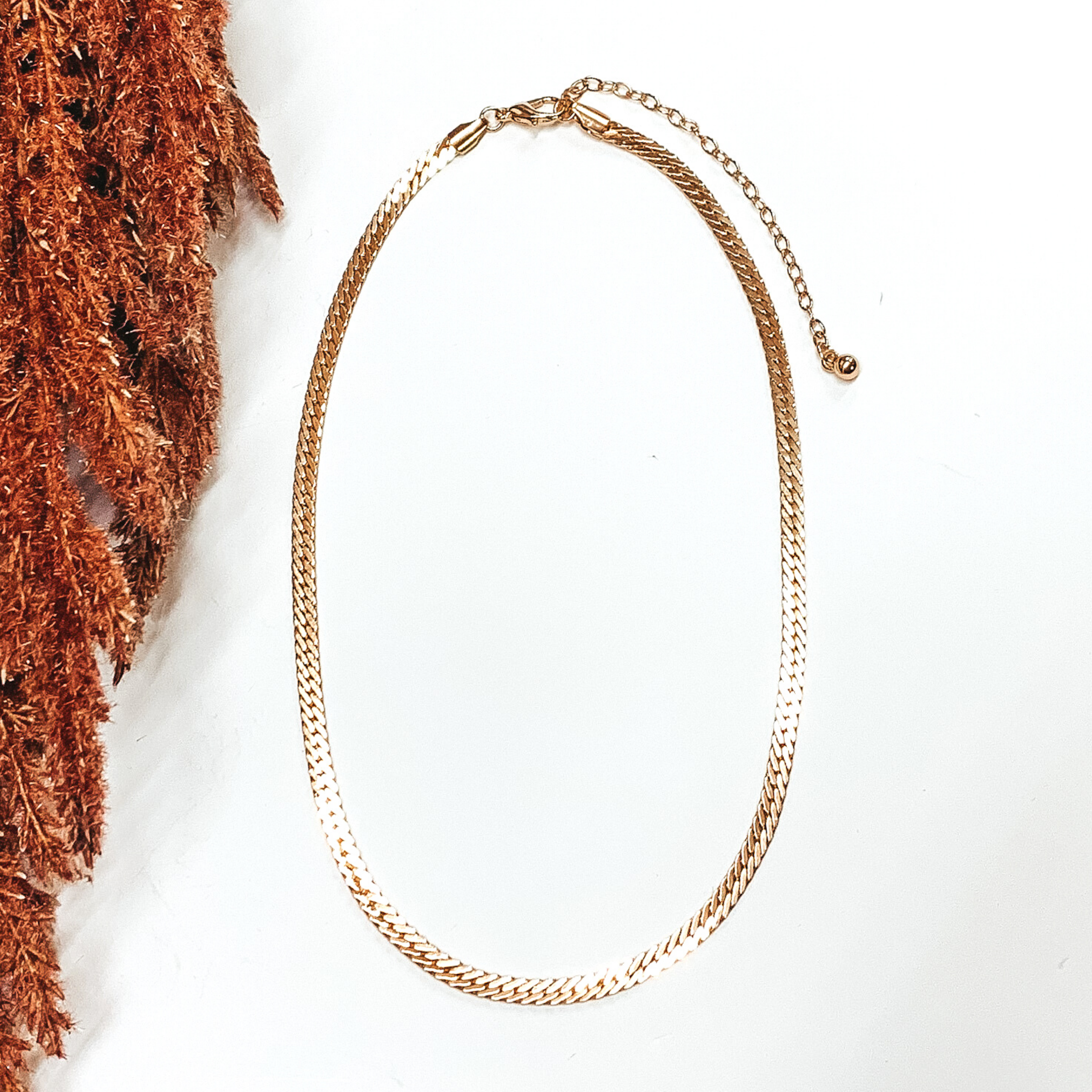 Simple flat snake chain necklace in gold. This necklace is pictured on a white background with brown floral on the side of the pic.