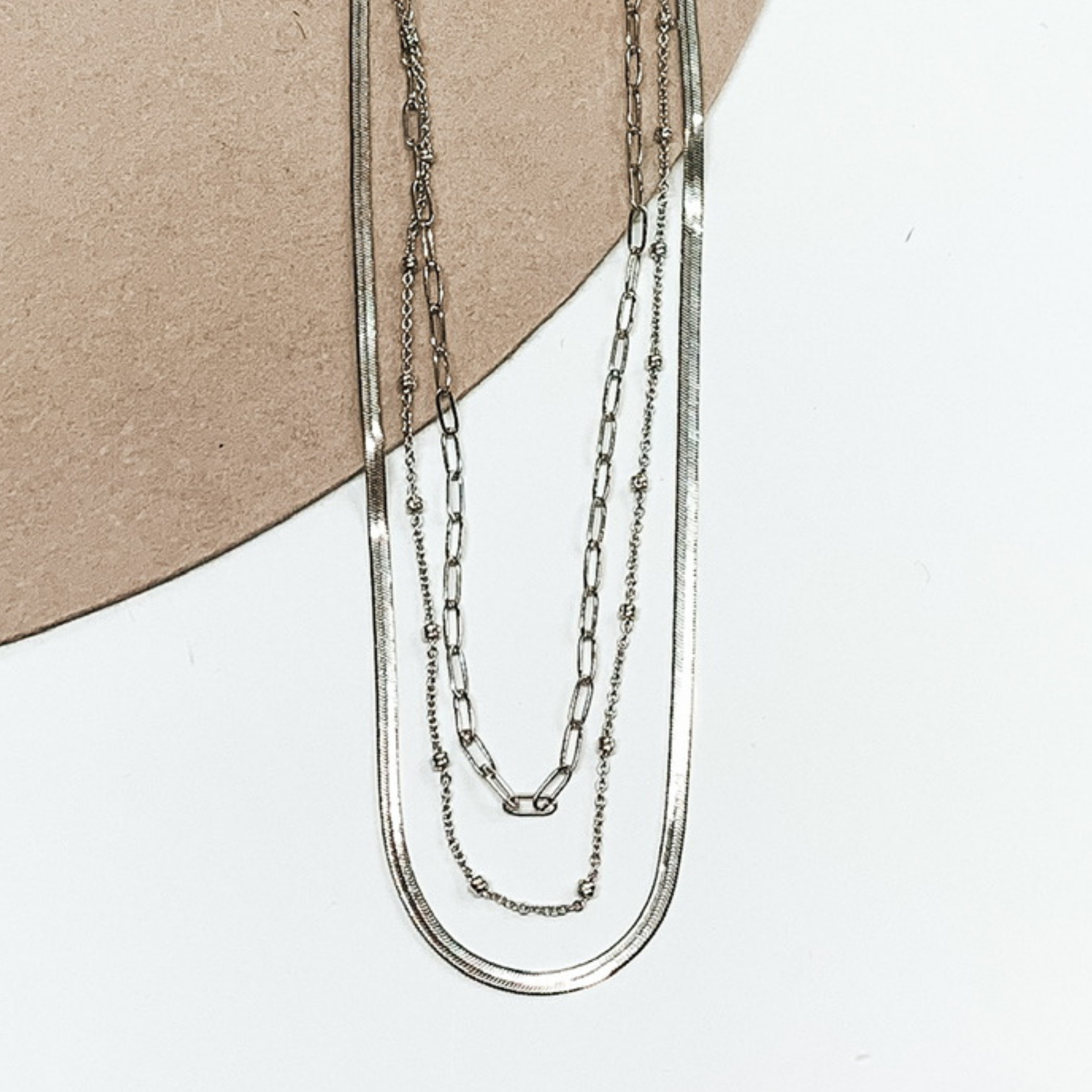 This silver necklace has three different chains. The first one is a classic thin link chain strand, the next one has tiny silver bead spacers, and the last one is a flat herringbone chain. This necklace is pictured on a white and beige background. 