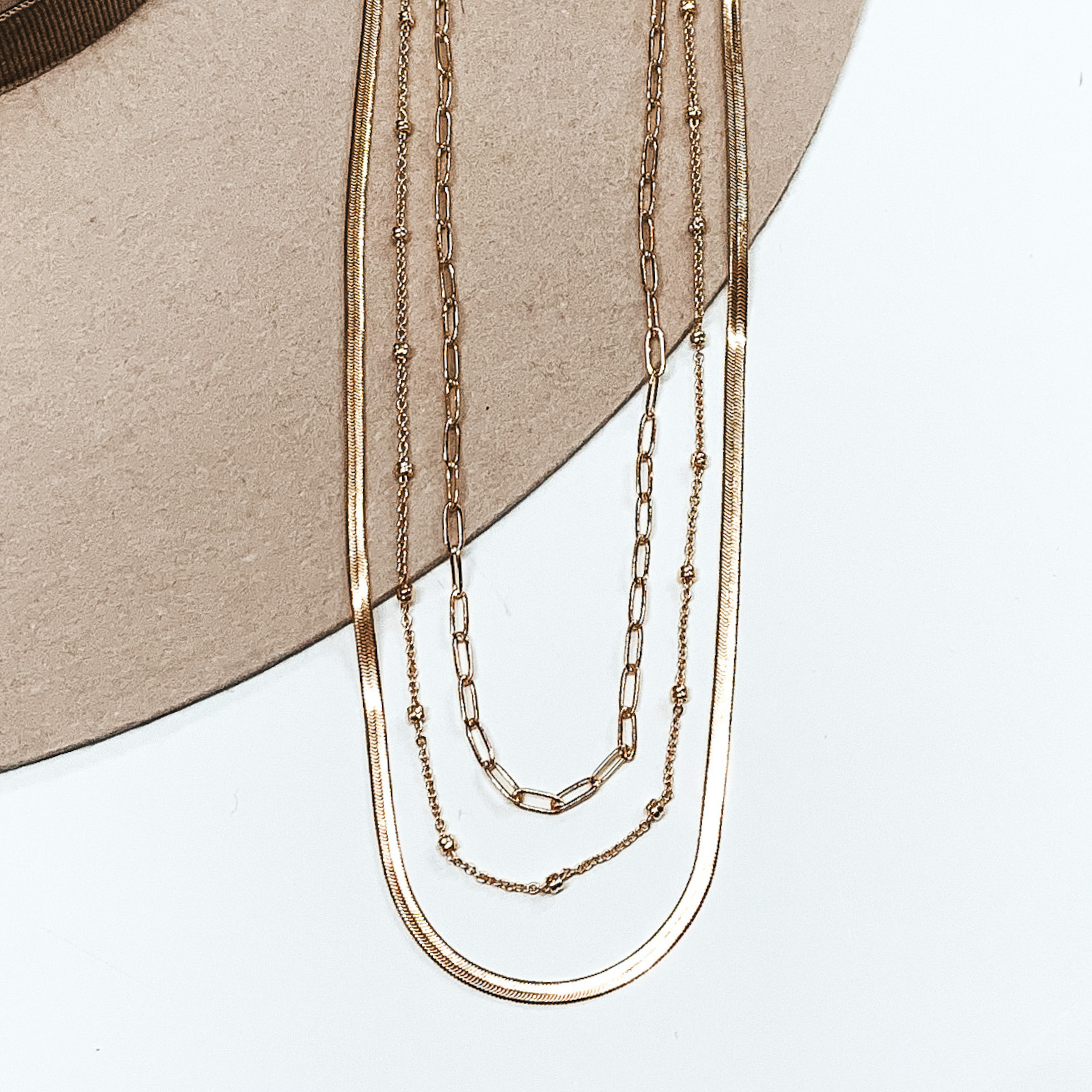 This gold necklace has three different chains. The first one is a classic thin link chain strand, the next one has tiny gold bead spacers, and the last one is a flat herringbone chain. This necklace is pictured on a white and beige background.