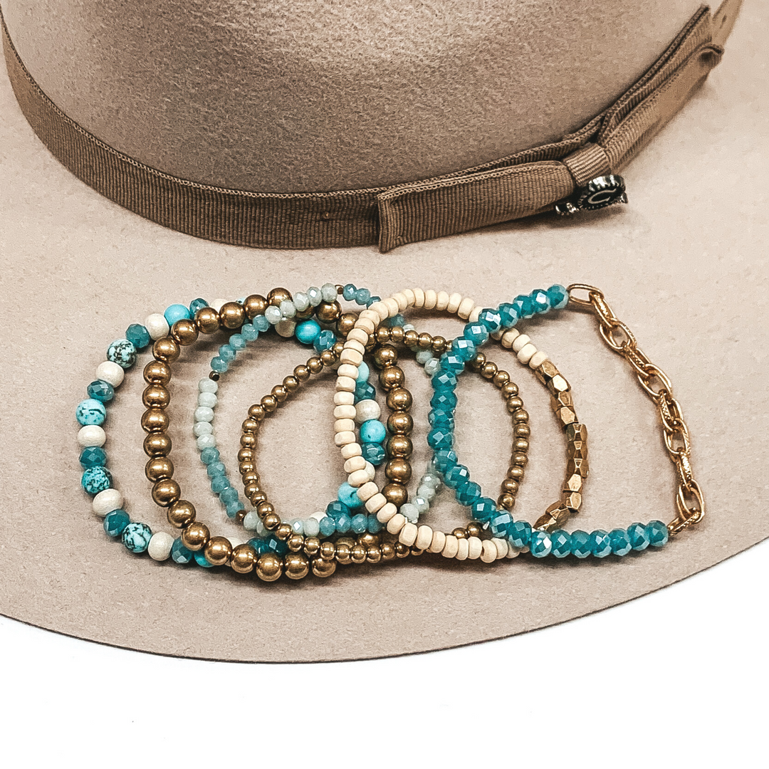 This bracelet set has 6 different beaded bracelets. One has different hues of teal beads, you have two gold beads (one is small and the other is bigger), one bracelet has two different hues of teal crystal beads that has gold spacers, one has teal crystal beads with a gold chain on one half of it, and the last bracelet has ivory and gold beads. This bracelet set is pictured on a beige hat on a white background.