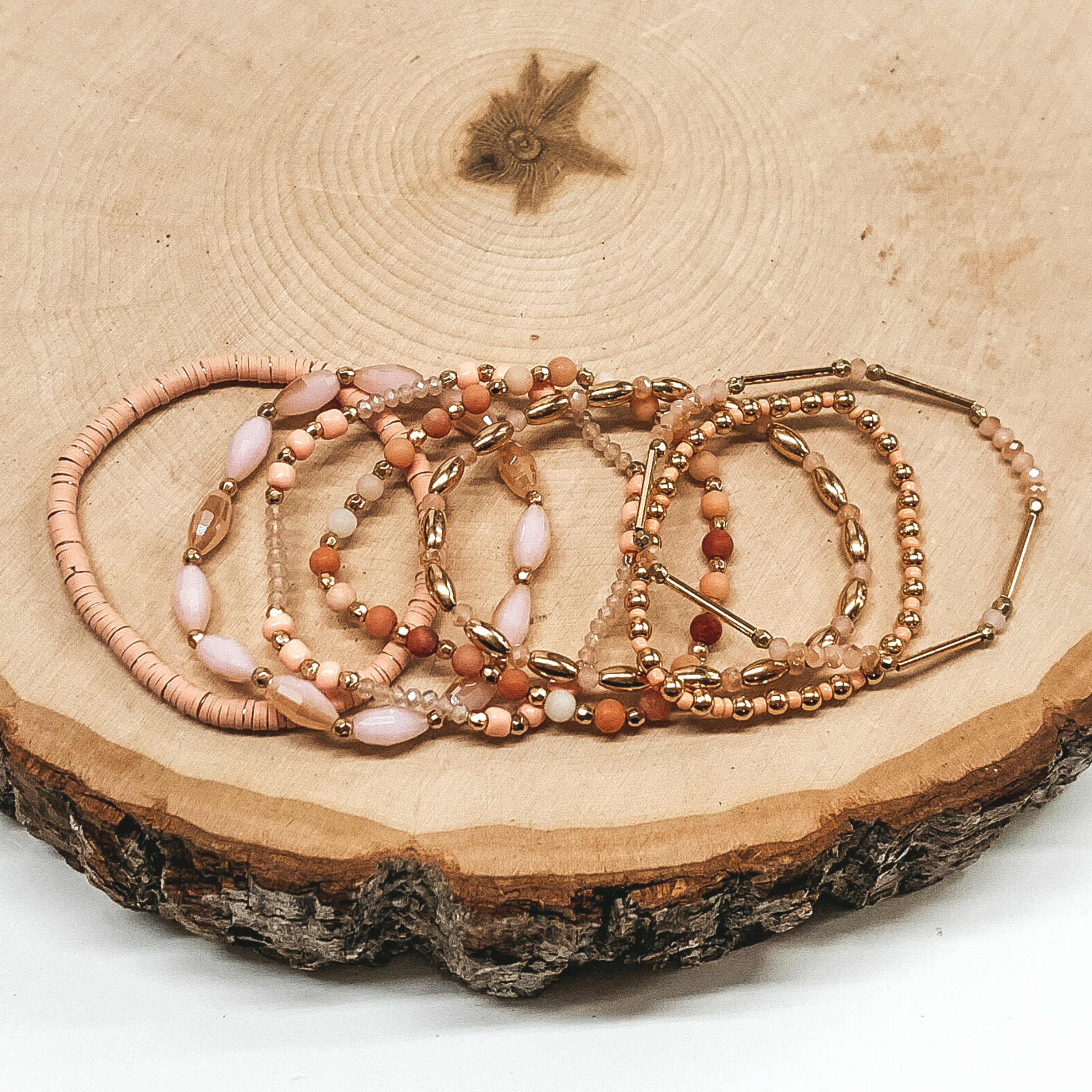 This bracelet set is a mix of light pink and gold beads. All seven bracelets are a different mix of beads, including, disk beads, crystal beads, circle beads, and bar beads. These are pictured on a piece of wood on a white background.