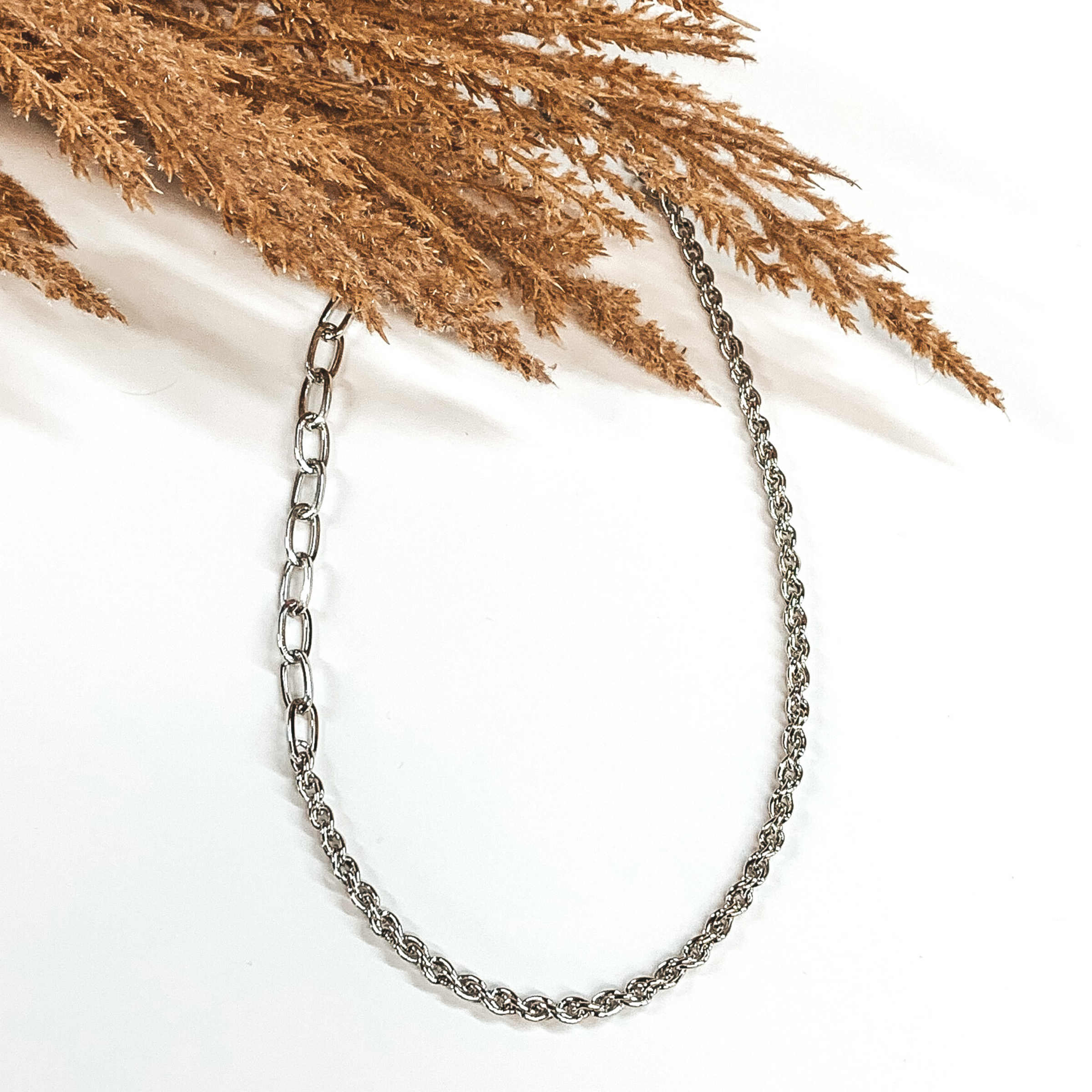 A single silver chained necklace. This necklace includes two different types of chains on the same strand. This necklace is pictured on a white background with tan floral at the top. 