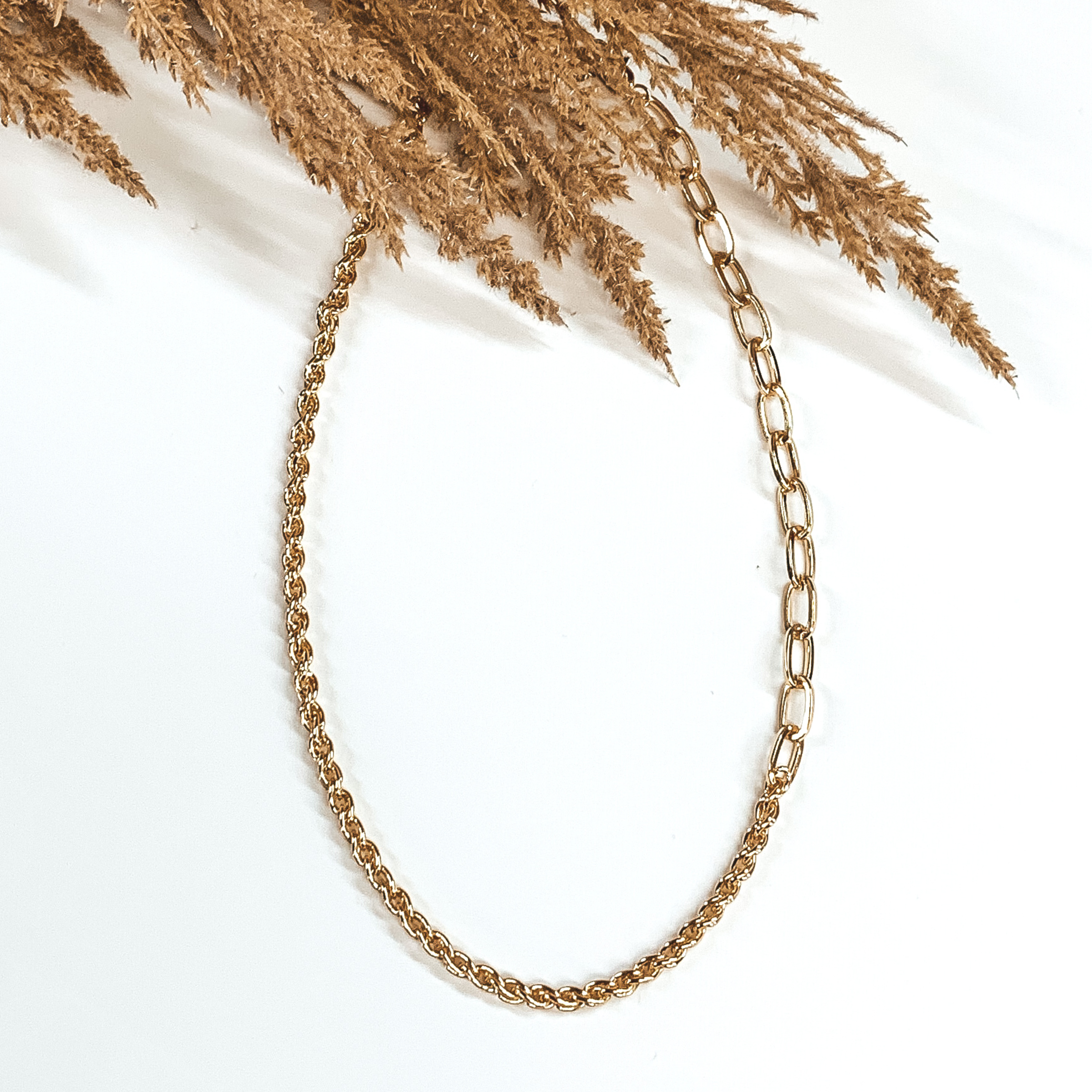 A single gold chained necklace. This necklace includes two different types of chains on the same strand. This necklace is pictured on a white background with tan floral at the top.