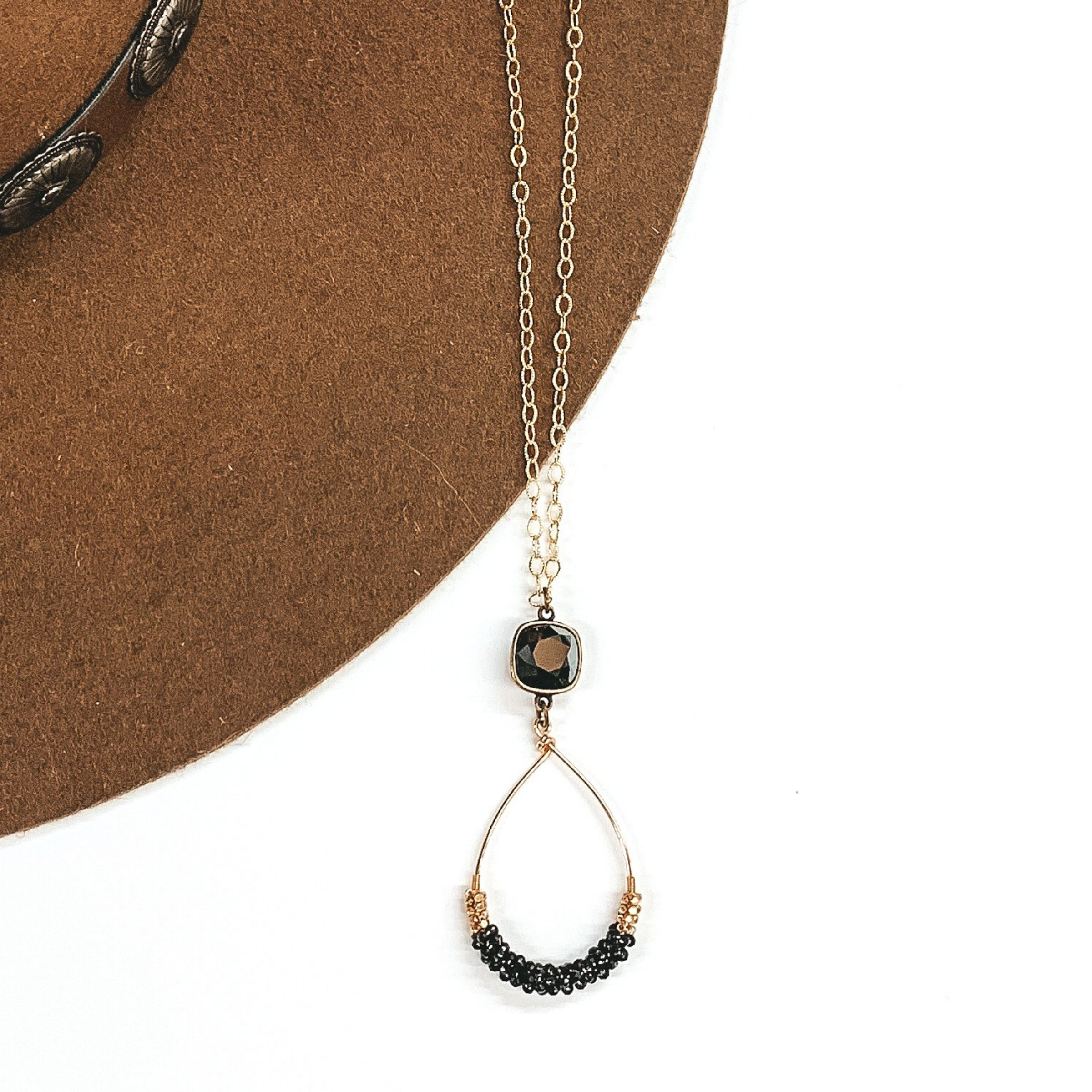 Gold chained necklace with black cushion cut crystal charm that has a hanging teardrop pendant with black and bold beads on the teardrop. This necklace is pictured on a brown and white background. 