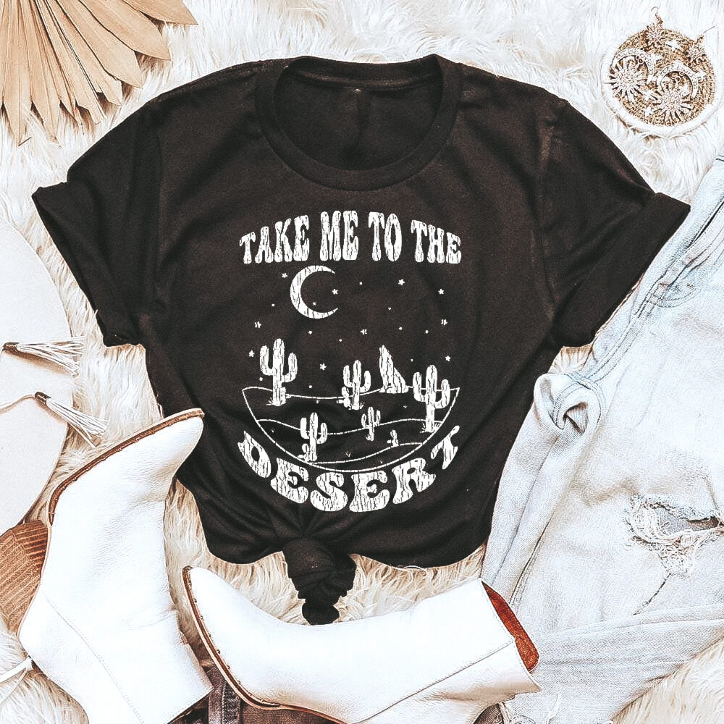Black graphic tee with "Take me to the desert" in cute font. Surrounded by stars, cacti, moon and coyote You can see this tee paired with white booties and light wash jeans. Shirt is laid on top of a fuzzy rug with cute trinkets surrounding.