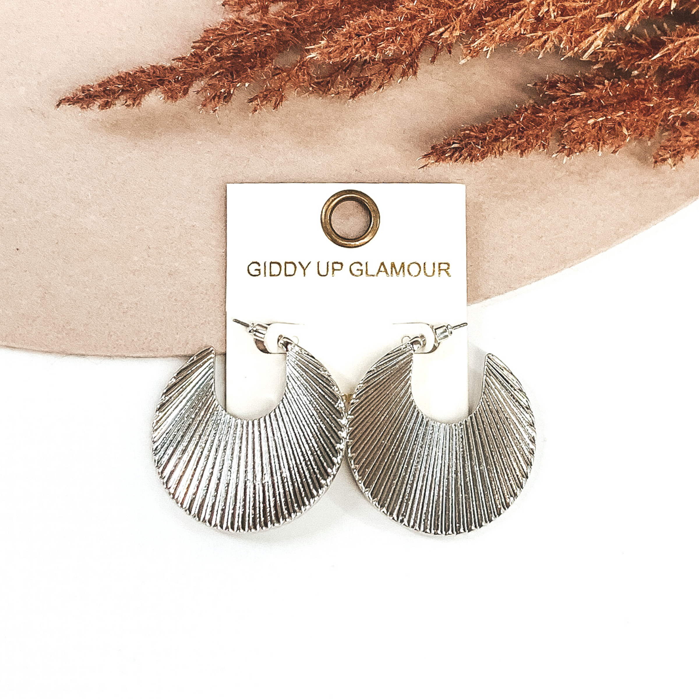 These silver earrings are an irregular curved shaped hoop earrings with notch at the top. These earrings are pictured on a jewelry back that is laying on a white and beige background with brown floral at the top. 